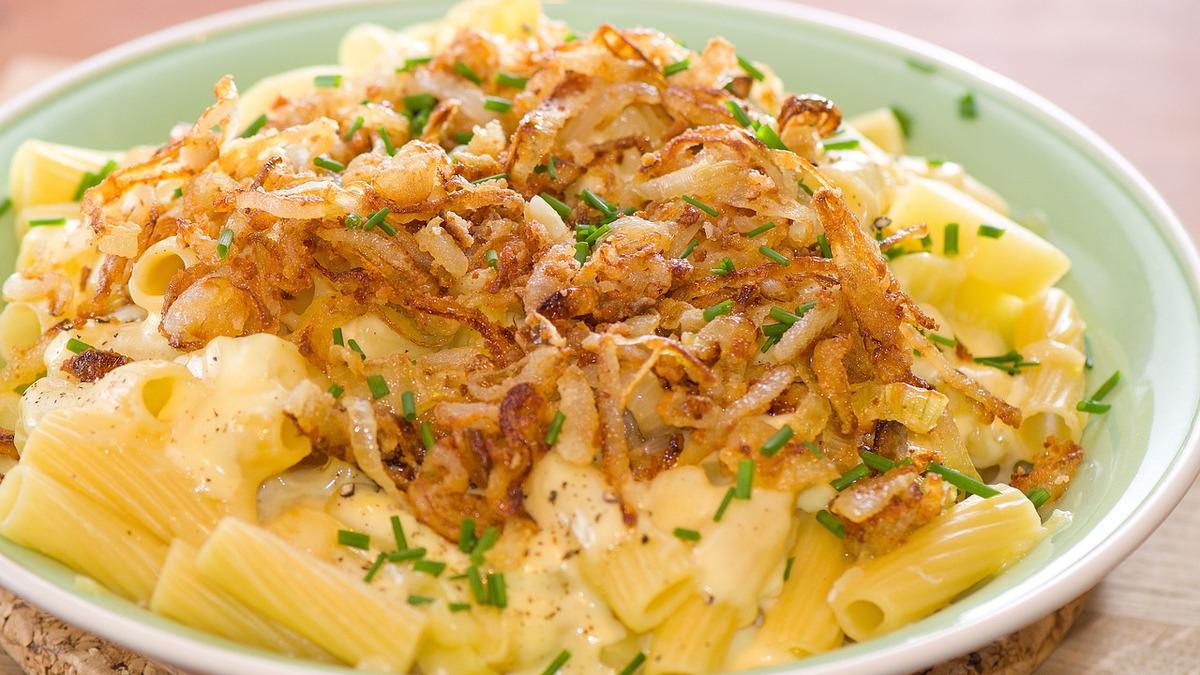 A bowl of rigatoni pasta is covered in a creamy sauce and topped with fried onions and chopped chives. The dish is served in a light green bowl.