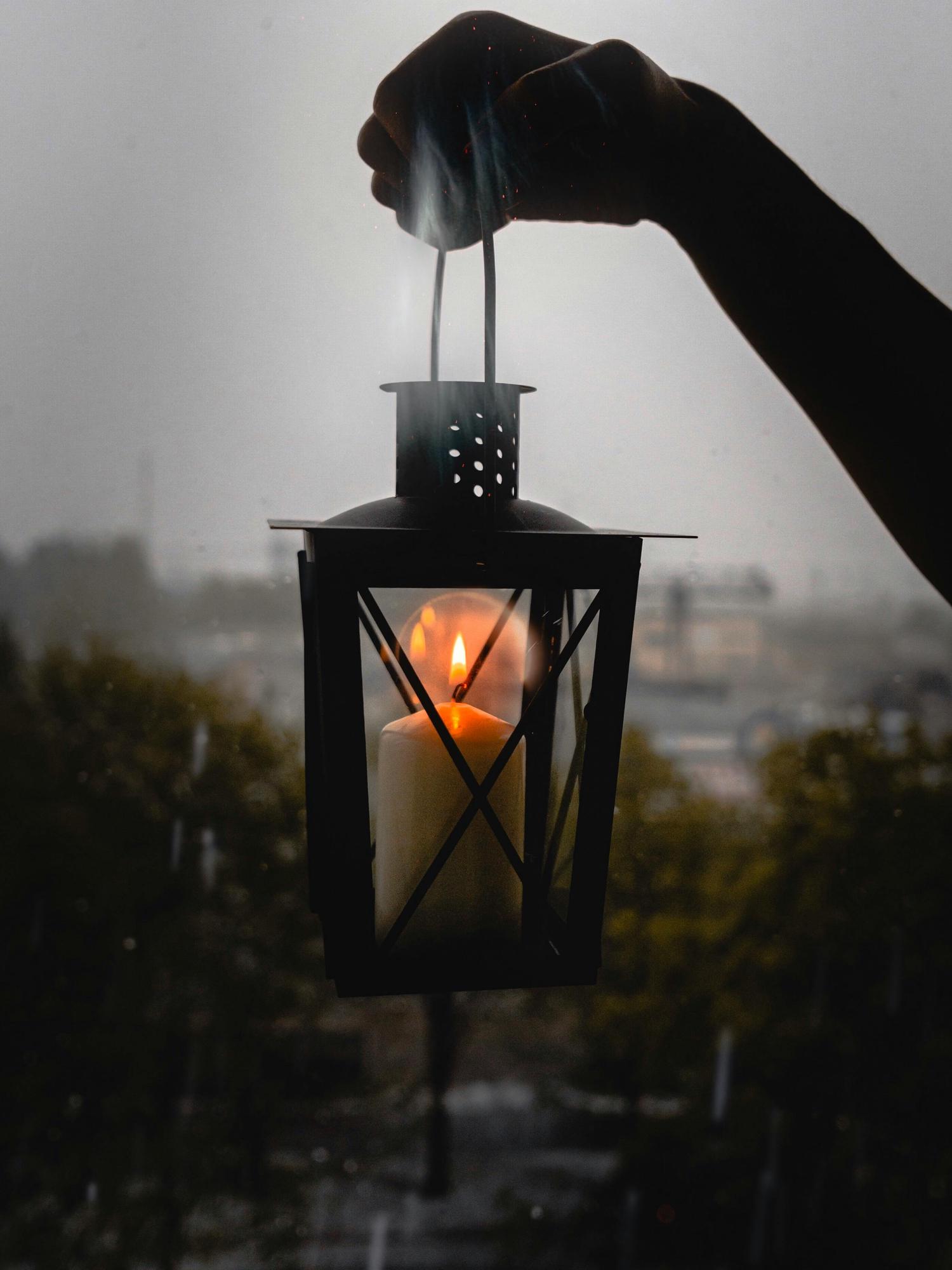 A silhouetted hand holds a black lantern with a lit candle inside, against a blurred background of a rainy cityscape. The candle flame glows brightly, casting light within the lantern.