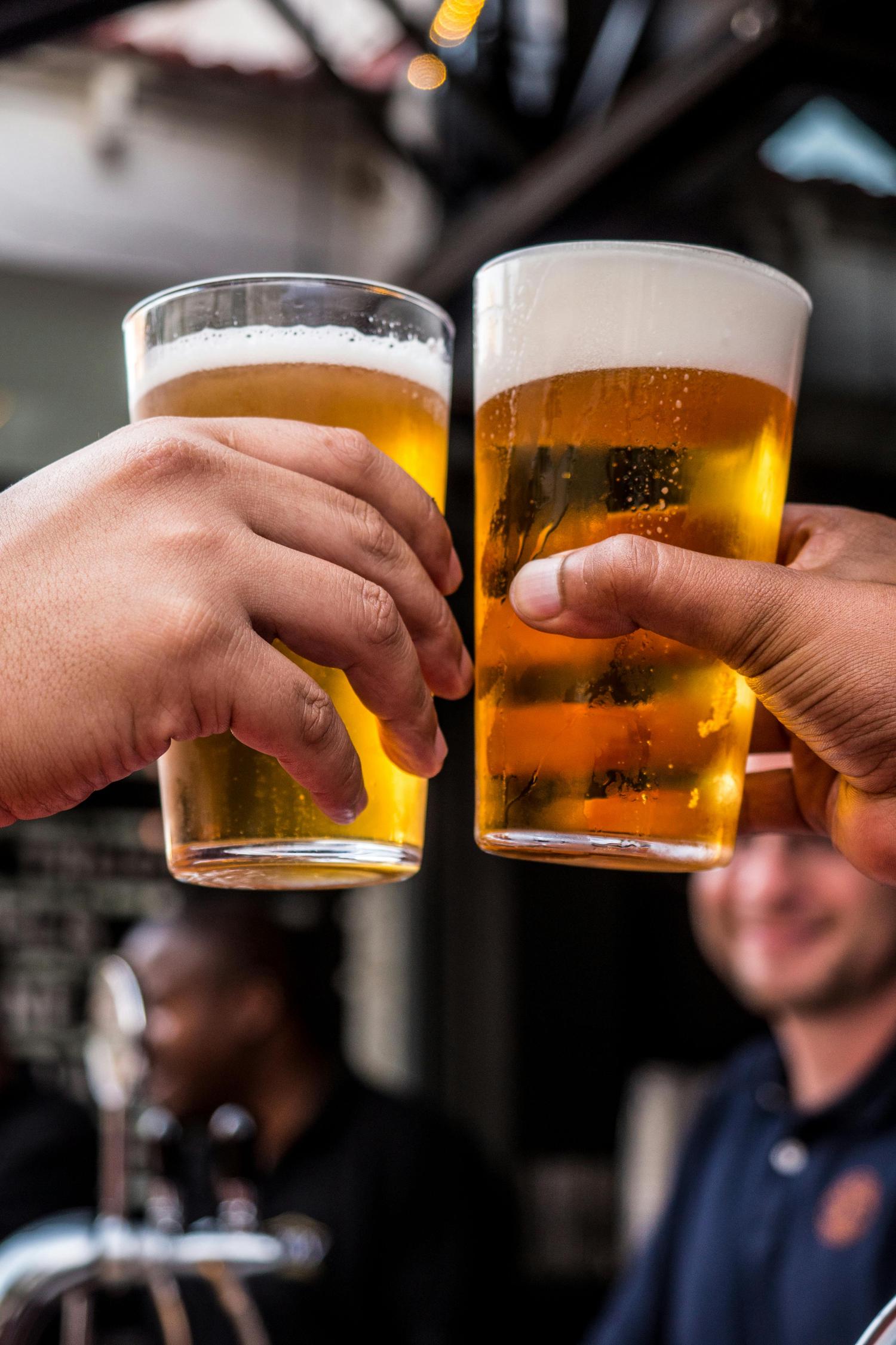 Two hands hold glasses of golden beer with foamy heads, clinking them together in a celebratory toast at an outdoor bar. In the blurred background are other patrons.
