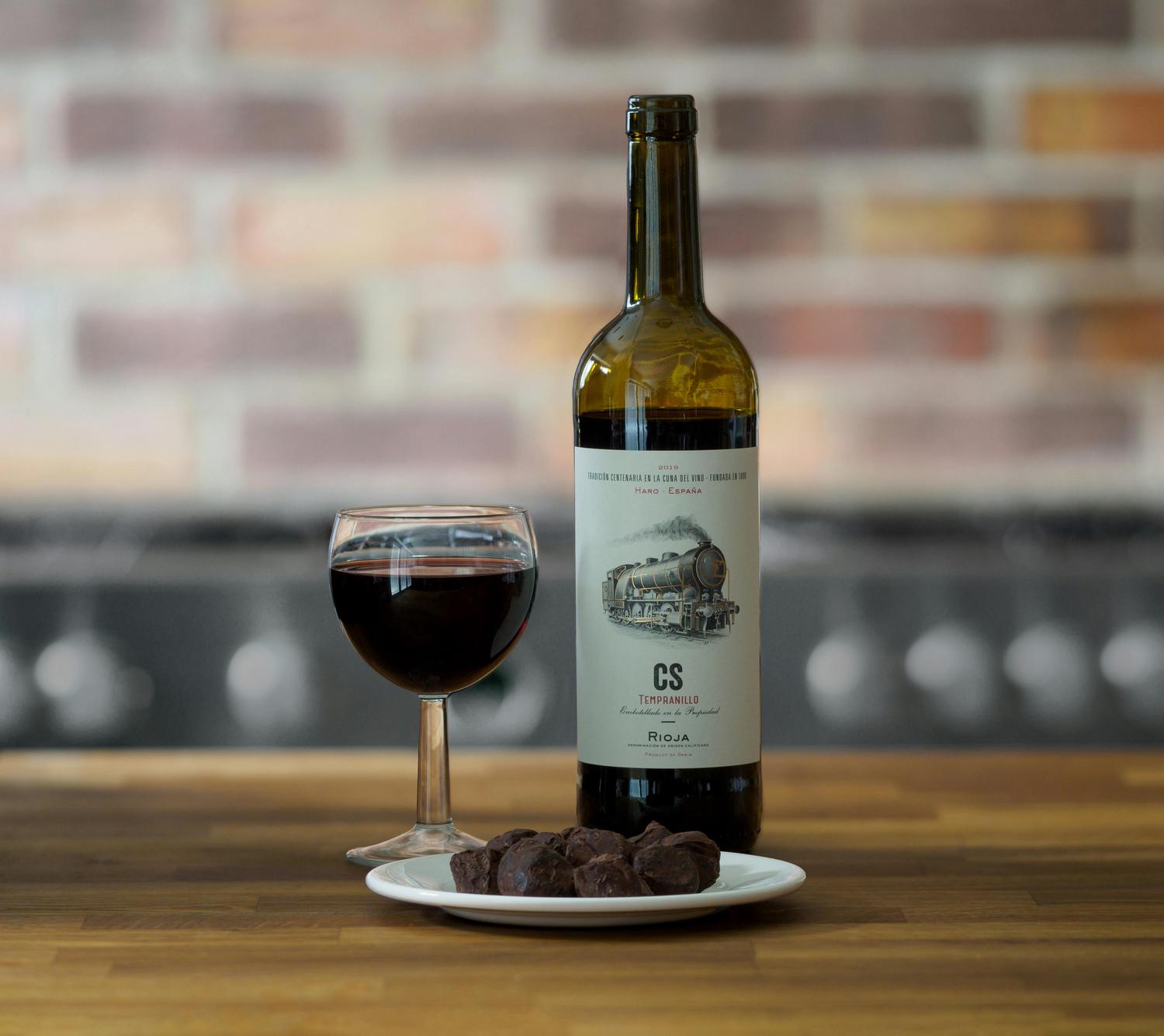 A bottle of red wine with a label depicting a steam train sits on a wooden counter, next to a wine glass filled with red wine and a plate of chocolate truffles. The wine bottle is labeled 