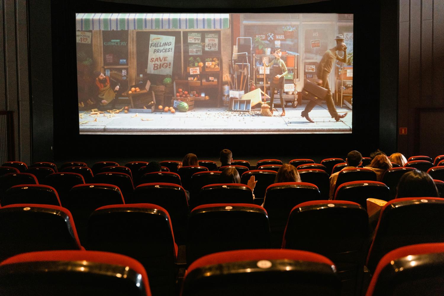 A movie theater showing an animated film on the screen above rows of seated viewers. The film shows a street scene with a grocery store and hardware store.