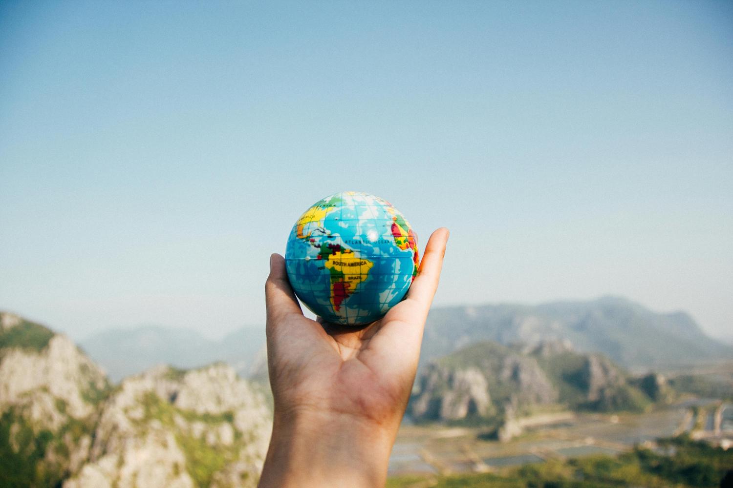 A hand holds a small, colorful globe against a backdrop of blurry mountains and a clear blue sky. The globe features visible continents and oceans.