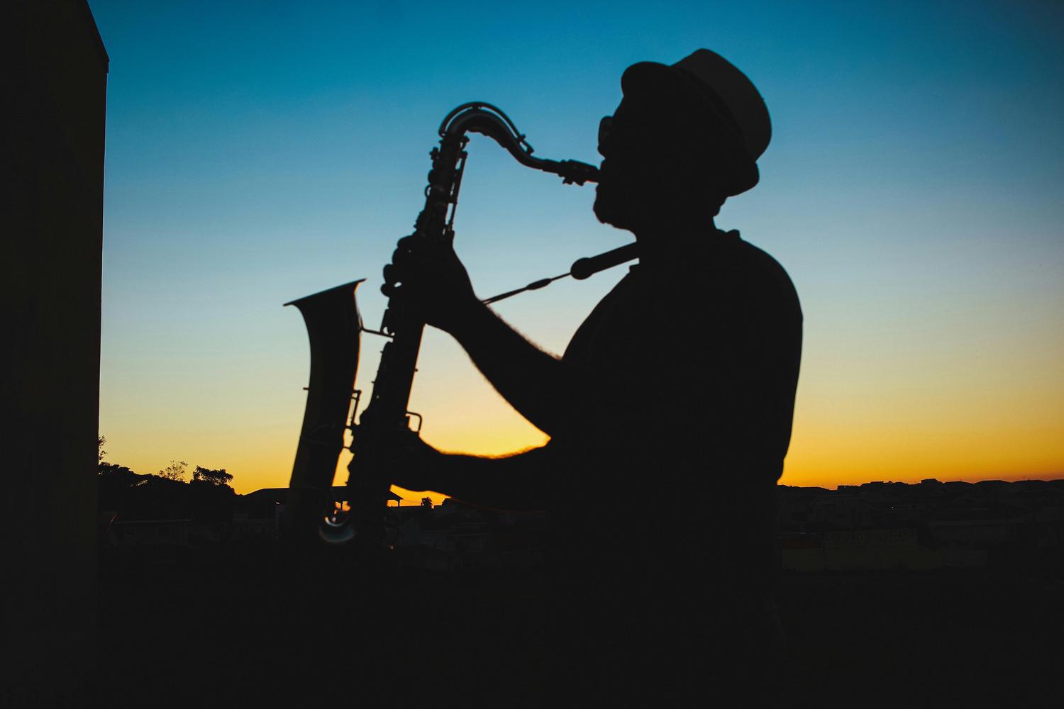 Silhouette of a person with a hat playing a saxophone against a vibrant sunset sky. Buildings are visible in the distance under the horizon line.