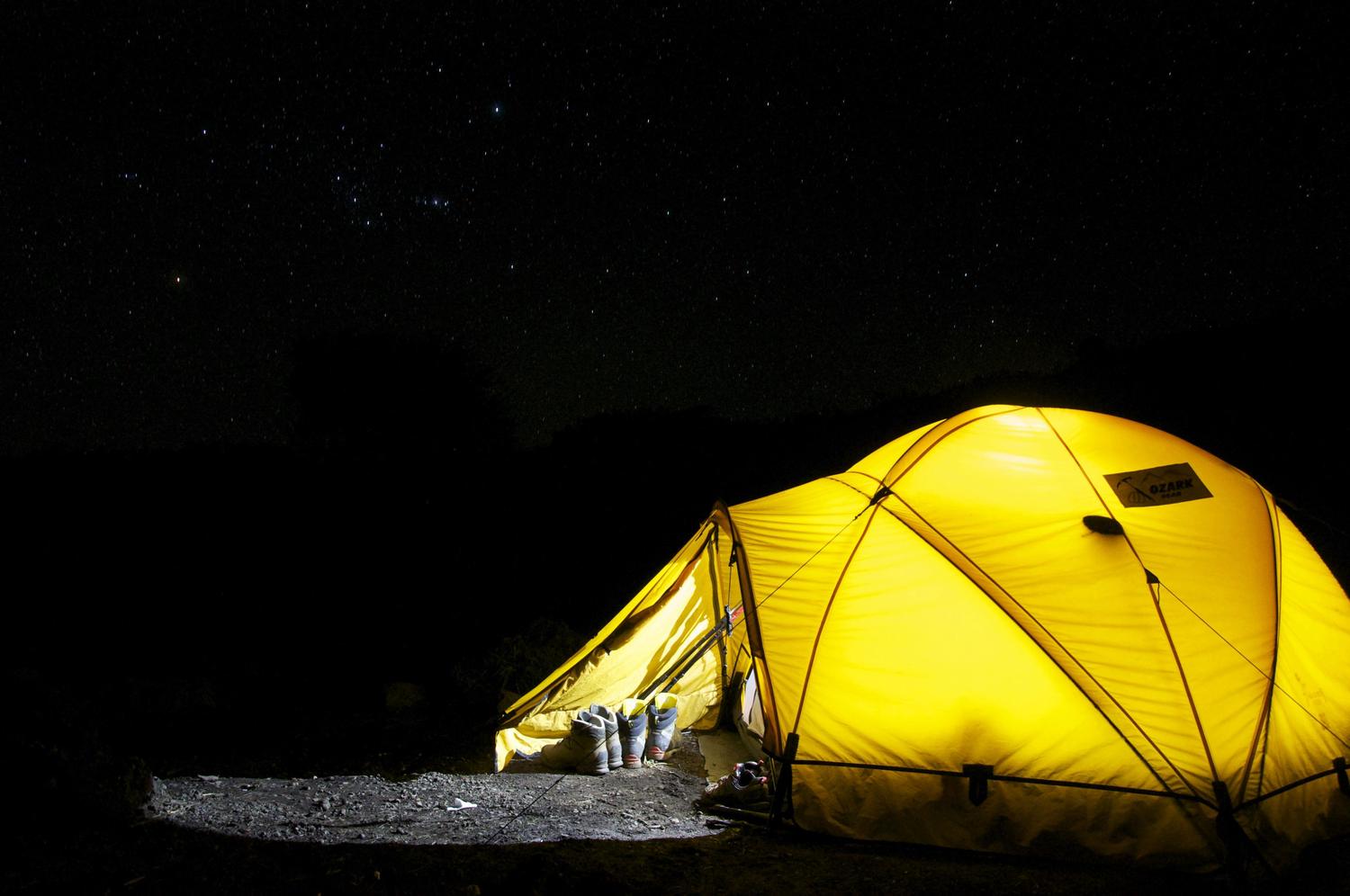 A bright yellow tent glows against a dark night sky dotted with stars. Three pairs of hiking boots sit outside the tent's open entrance.