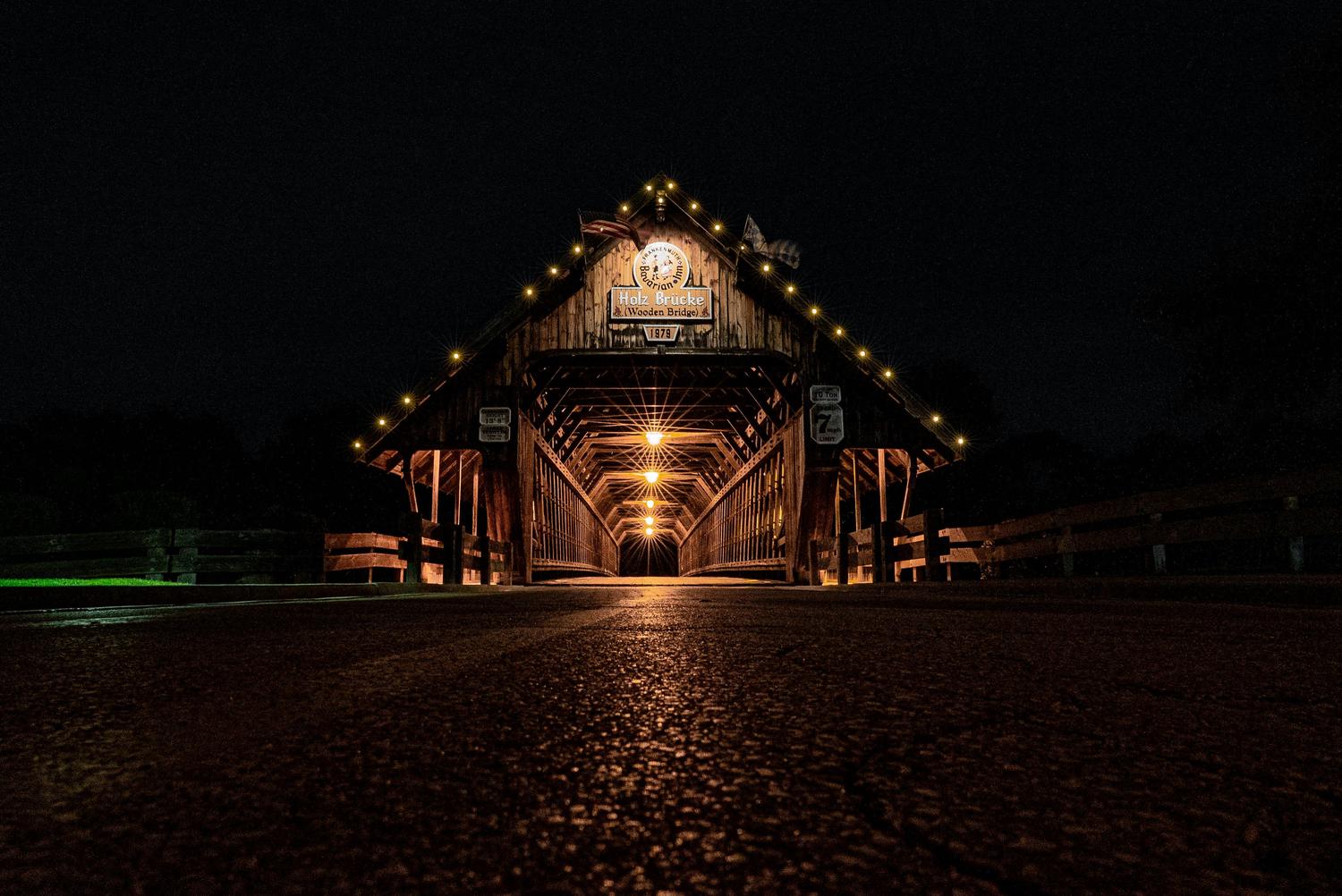 A low-angle shot at night shows the Holz Brucke wooden bridge illuminated by interior lights and decorative string lights. The bridge's entrance features signage in German and English, dating it to 1979, and also displays an American flag.