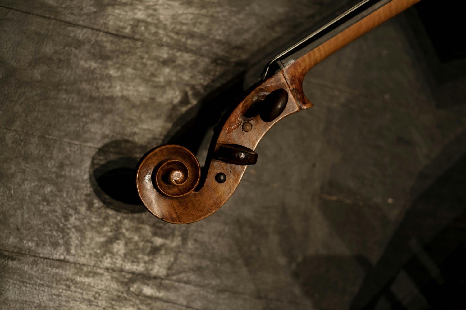 Close-up of a violin headstock with a decorative spiral end, strings, and tuning pegs, resting on a textured, dark grey surface. The wood shows signs of wear and age.