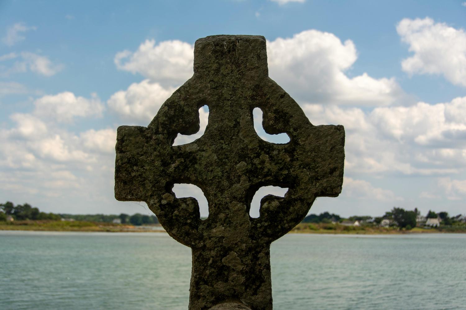 A weathered stone Celtic cross stands against a background of calm water, a distant shoreline with trees and houses, and a partly cloudy sky. The cross has carved openings at each arm's intersection with the center.