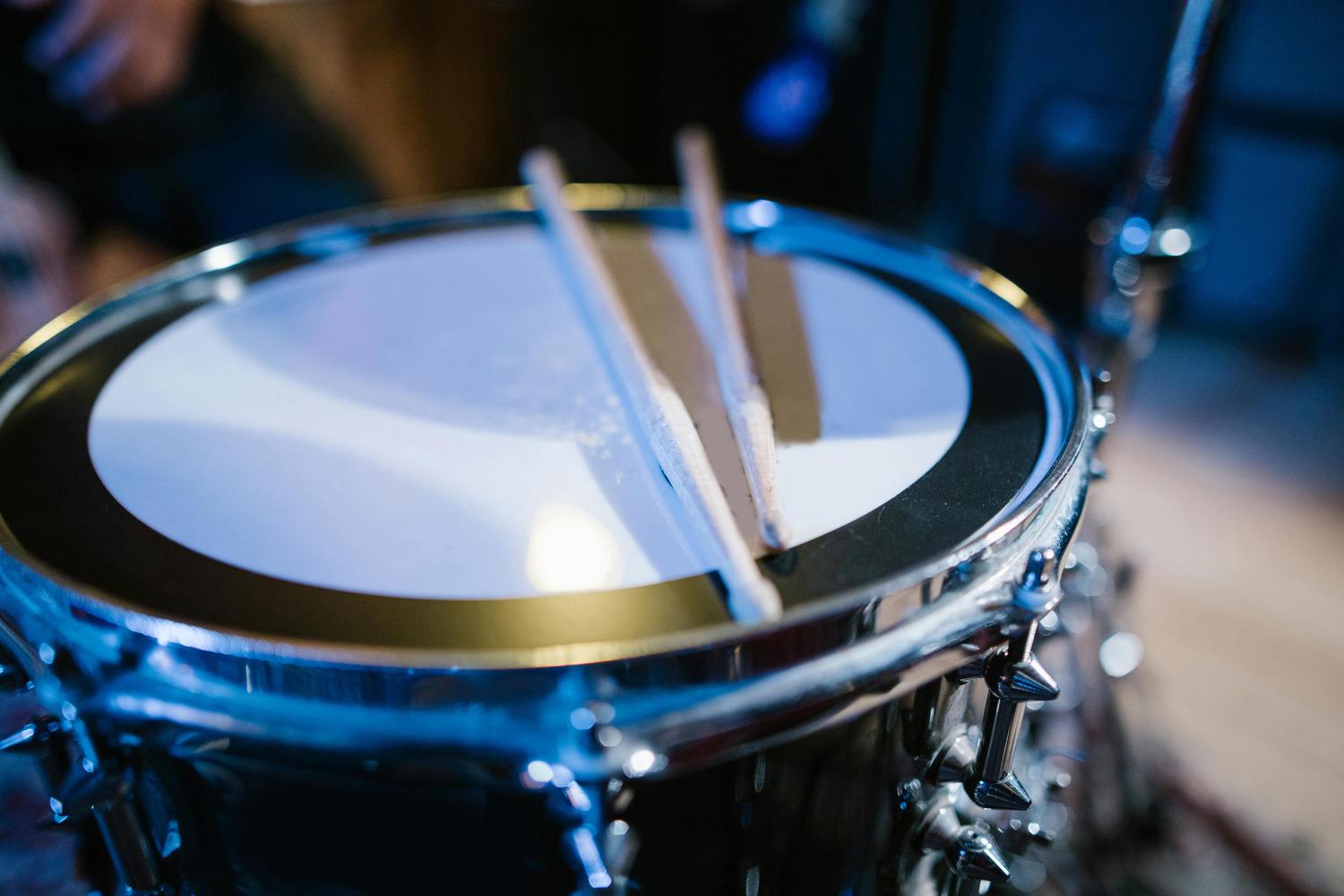 Close-up of a snare drum with two wooden drumsticks resting on its white head. The drum has a chrome finish with detailed tuning lugs.