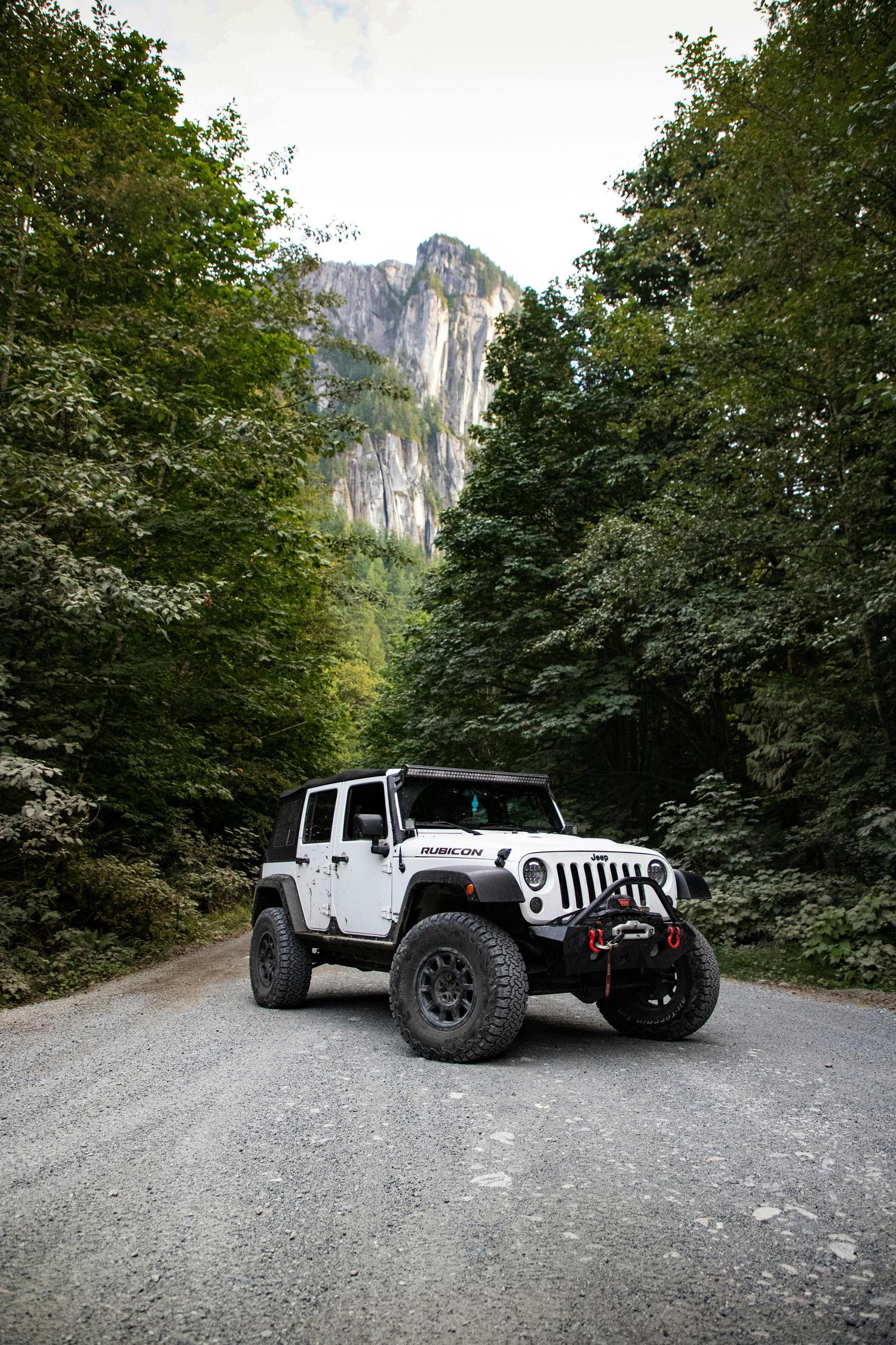A white Jeep Rubicon, equipped with off-road modifications, sits on a gravel road flanked by lush green trees with a towering rock formation visible in the distance. The scene suggests an outdoor adventure setting.