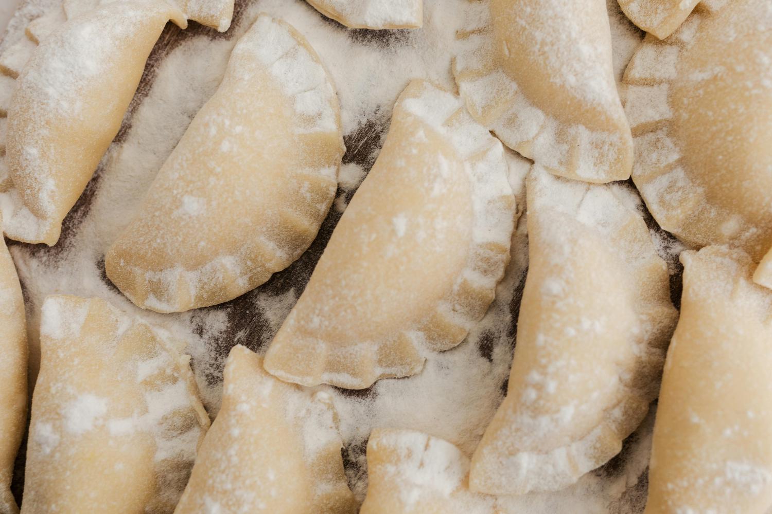 Close-up of uncooked pierogi sprinkled with flour, covering a dark surface. The pierogi are a pale beige color and have a crimped edge.