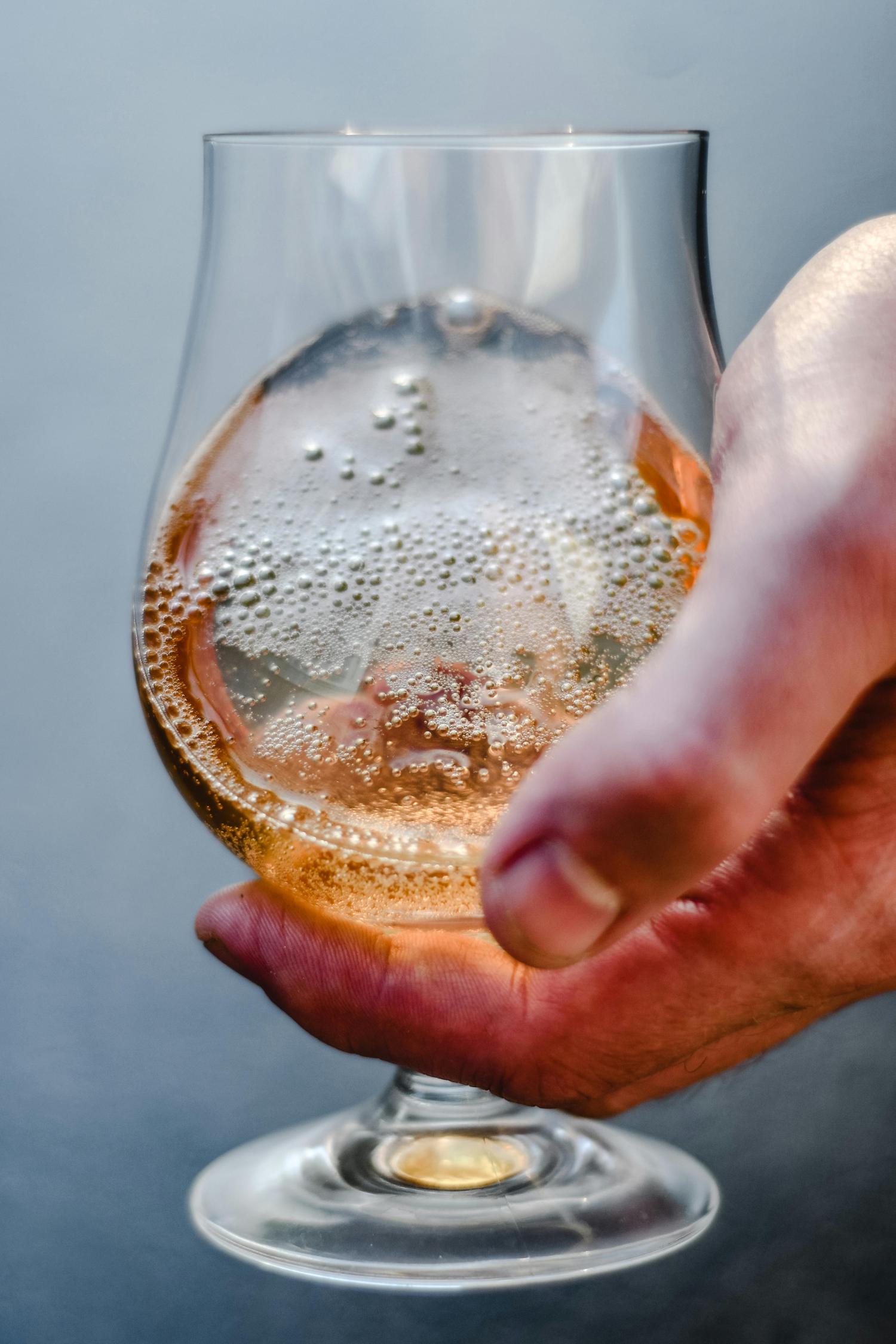 A hand holds a stemmed glass filled with a fizzy, golden-colored beverage, like cider or sparkling wine, against a blurred grey background. Bubbles cling to the sides and top of the drink.