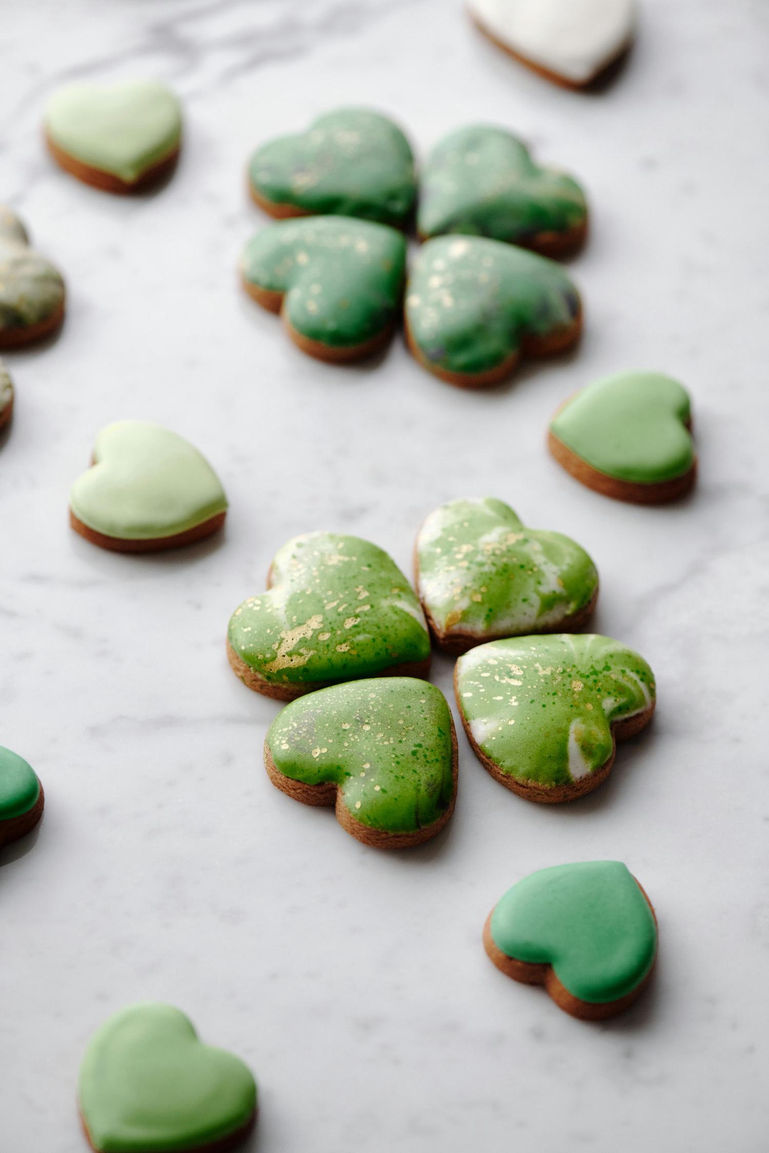 Heart-shaped cookies with green frosting and golden accents are scattered across a white marble surface. Some cookies are arranged in groups of four, resembling shamrocks.