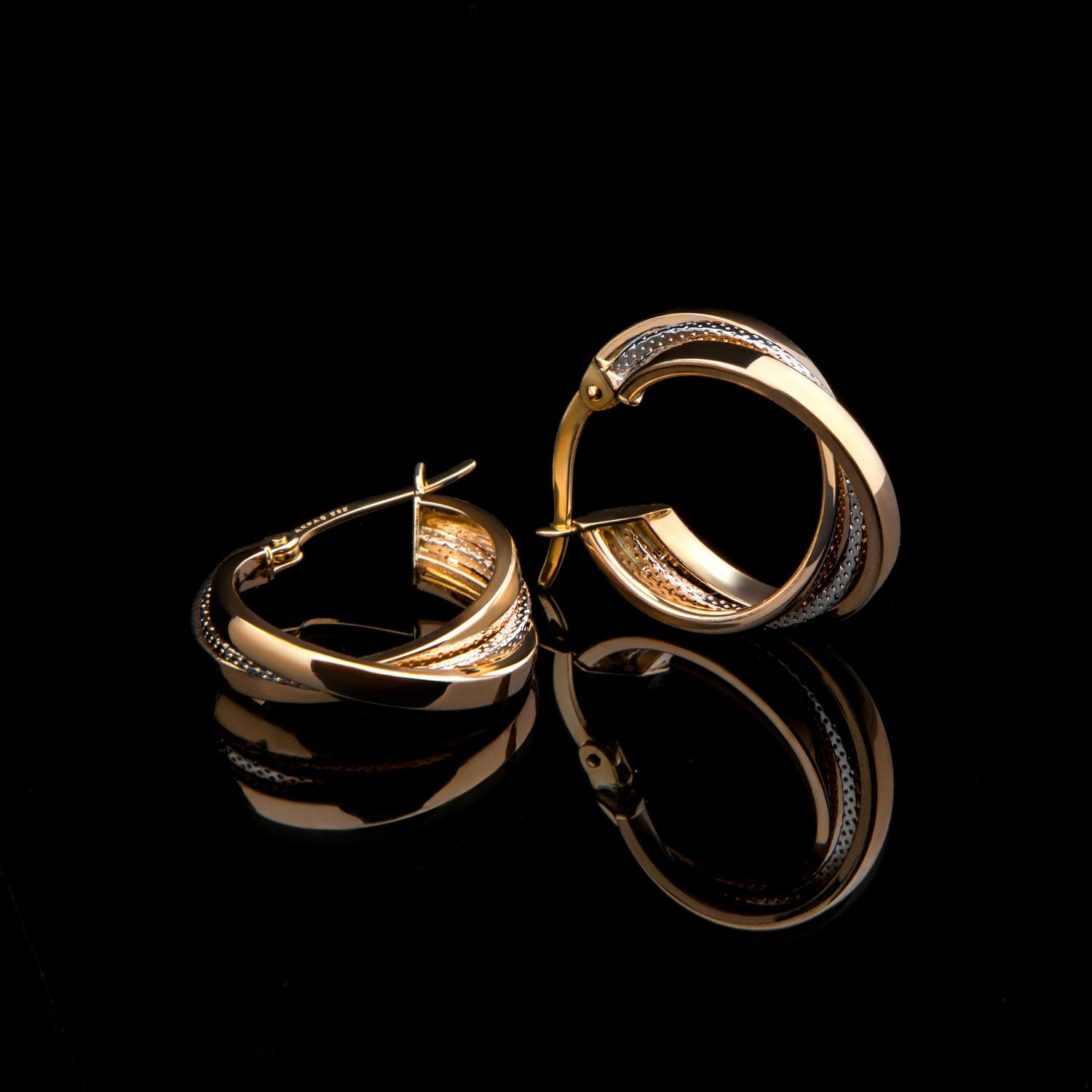 Two gold hoop earrings with a textured silver band sit on a glossy black surface, reflecting their shape. The earrings are presented in a way that showcases their design and shine.