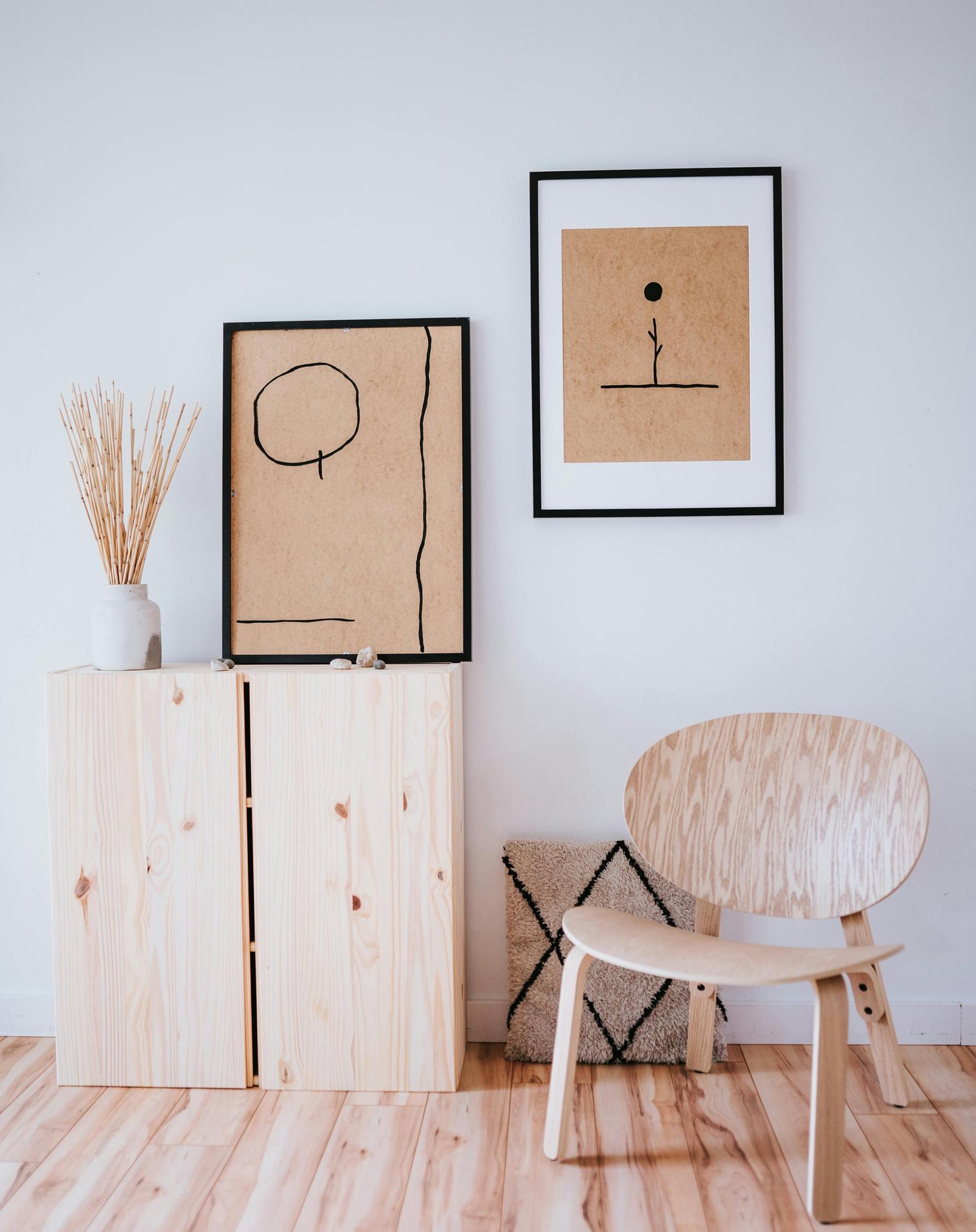 A minimal, modern room setting features a light wooden cabinet topped with a vase of dried stems and abstract framed art, along with a light wooden chair and textured rug in the corner against a white wall. The style is simple and clean, emphasizing natural materials and muted tones.