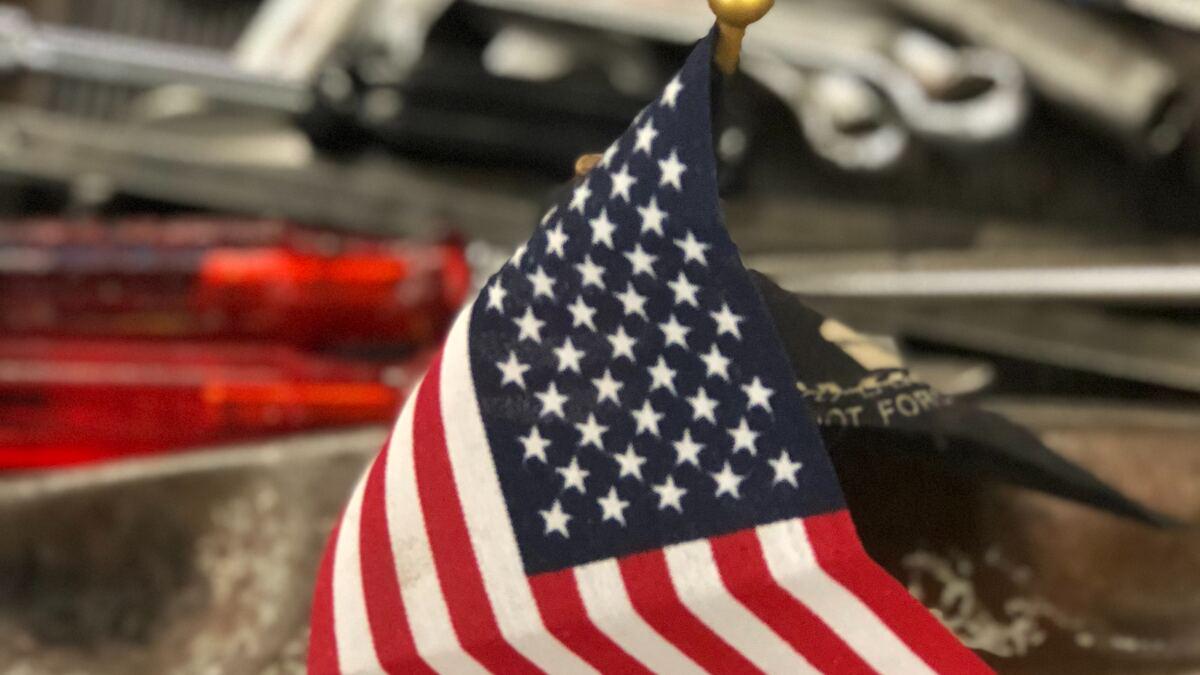 Close-up of a small American flag with a gold finial, with blurred tools and equipment in the background. A black baseball cap with white lettering is partially visible behind the flag.