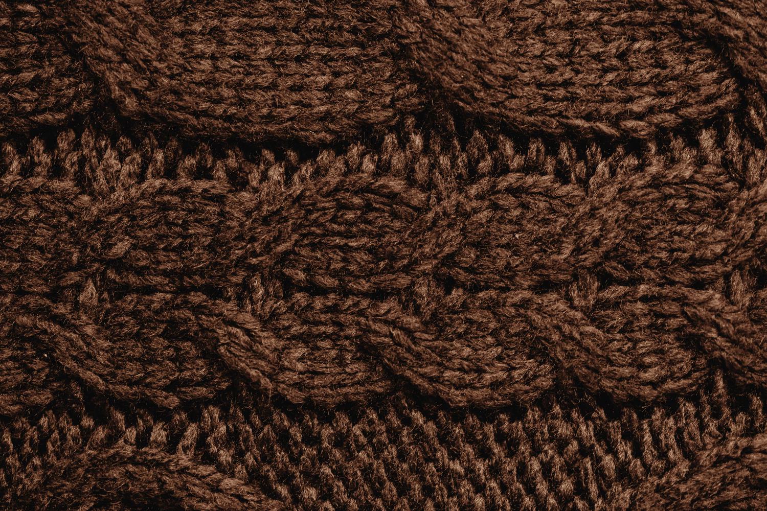 Close-up of brown knitted fabric with a cable pattern, creating a textured, warm background. The detail highlights the looped and interwoven threads of the yarn.