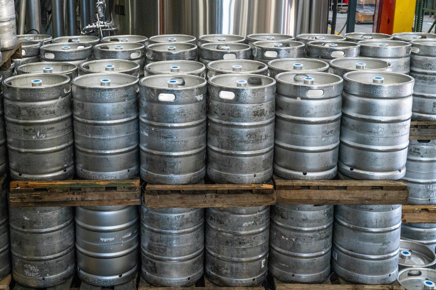 Stacks of Anheuser-Busch beer kegs are piled on wooden pallets in a brewery or warehouse. The metal kegs are cylindrical and display the Anheuser-Busch logo.
