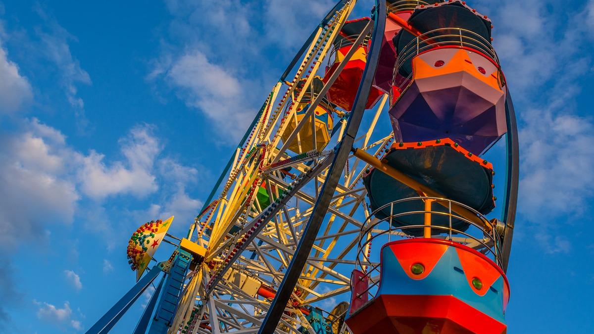 A low-angle shot of a brightly colored Ferris wheel against a blue sky with wispy clouds. The gondolas are decorated with geometric patterns in various colors.