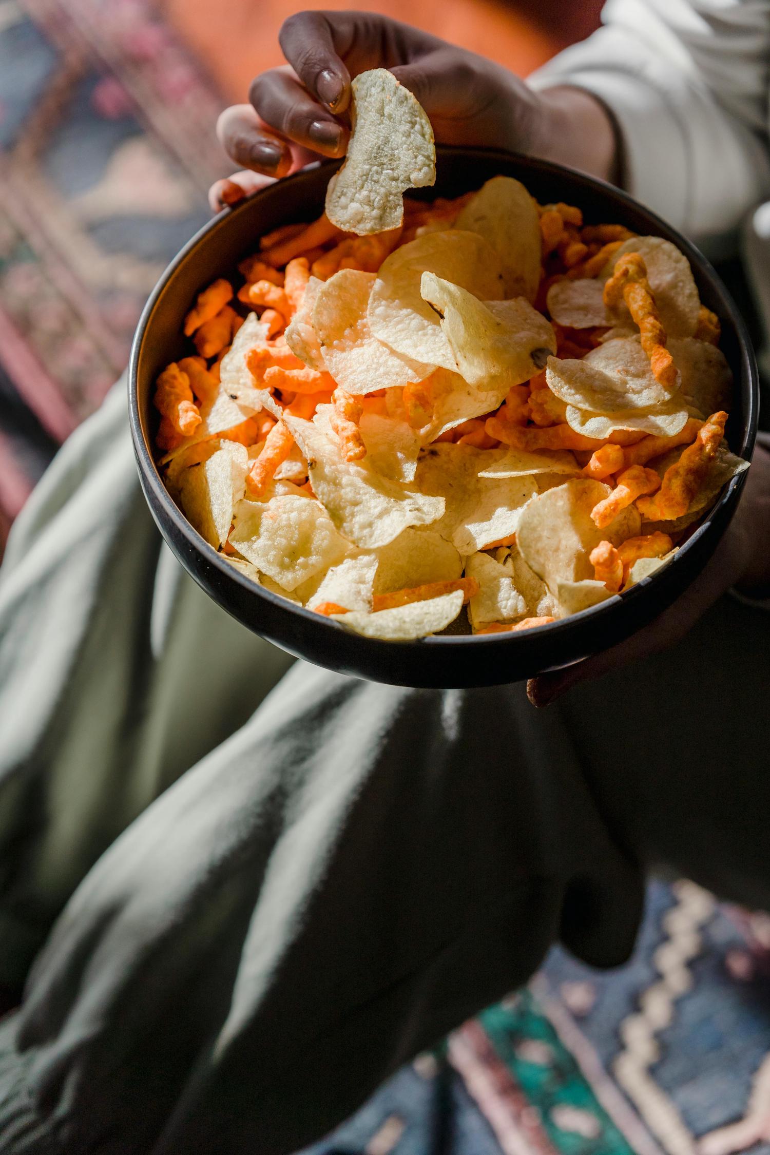 Hands hold a bowl filled with potato chips and cheese puffs. One hand reaches into the bowl to grab a chip.