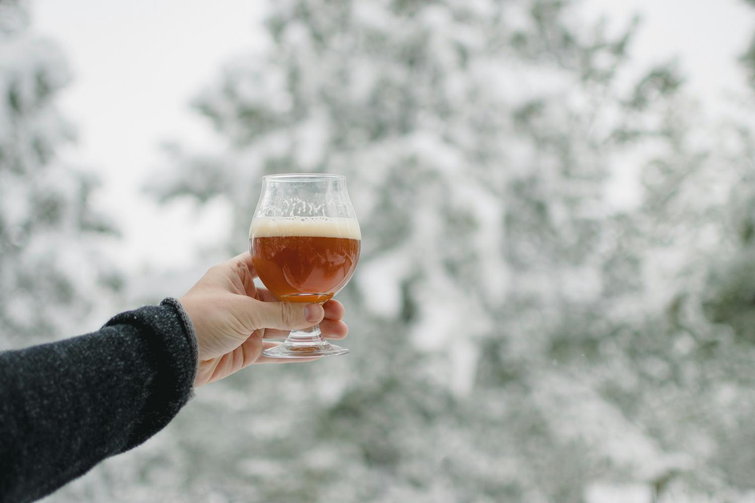 A hand in a dark sweater holds a tulip-shaped glass of amber-colored beer with a foamy head, against a blurry background of snow-covered trees. The scene suggests a wintery setting for enjoying a craft beer.