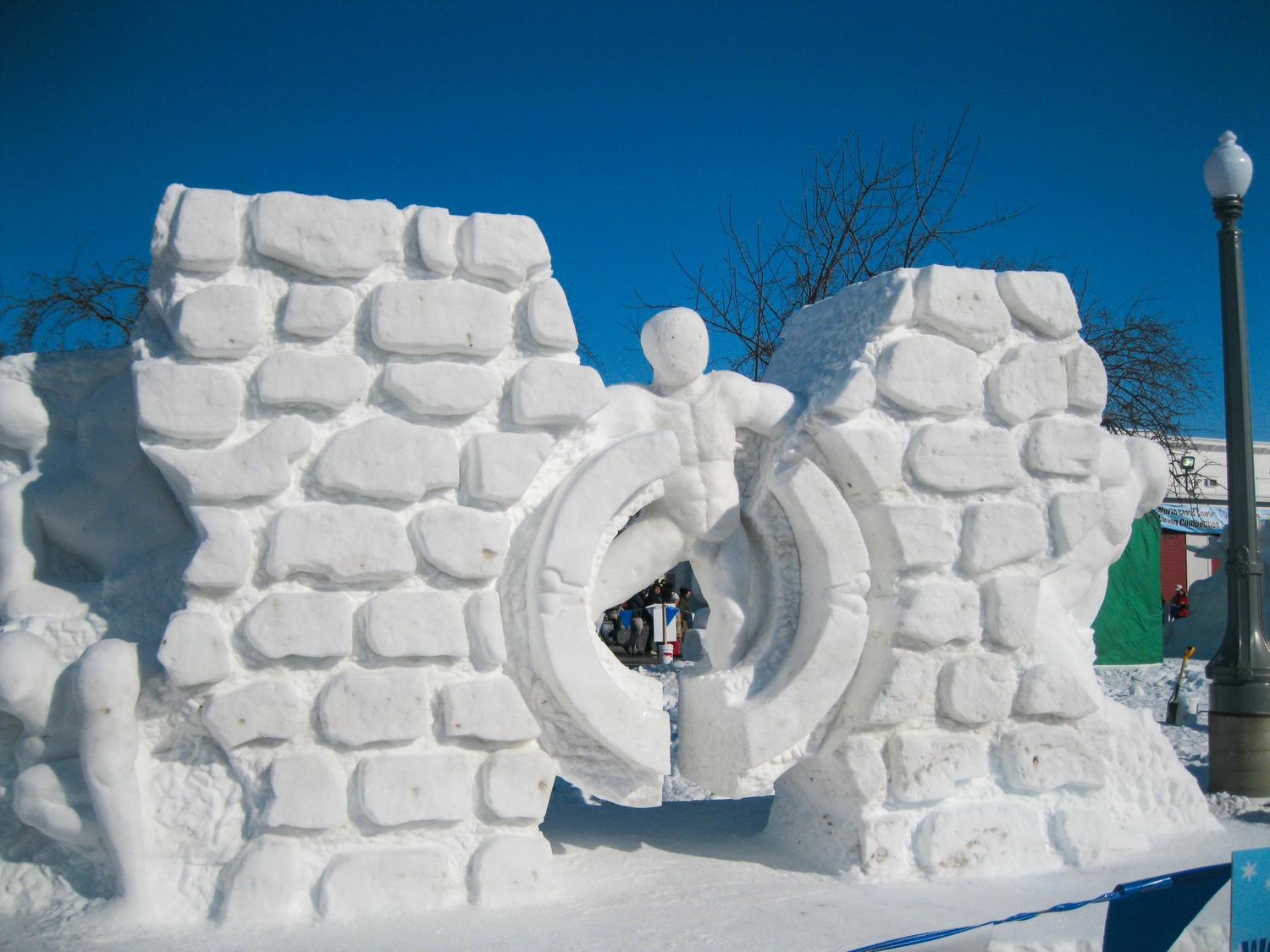 A snow sculpture of a figure pushing through a broken circular barrier set into a brick-like wall, under a clear blue sky. The sculpture is part of an outdoor event with a lamppost and other people visible in the background.