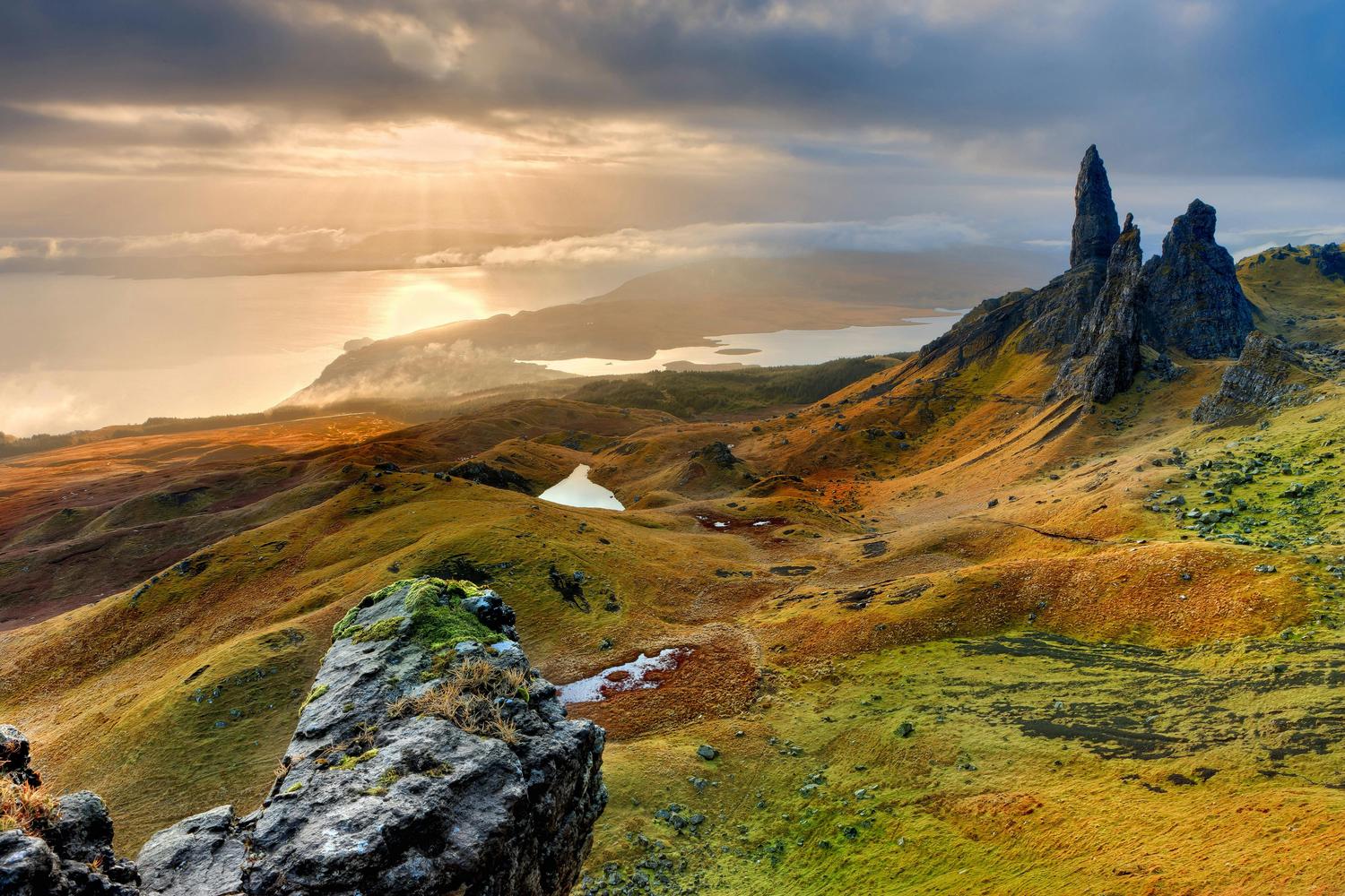 The Old Man of Storr, a tall rock pinnacle, rises above a rugged, hilly landscape with a loch and ocean visible in the distance under a sky with streaming sunlight. The foreground includes a moss-covered rock formation.