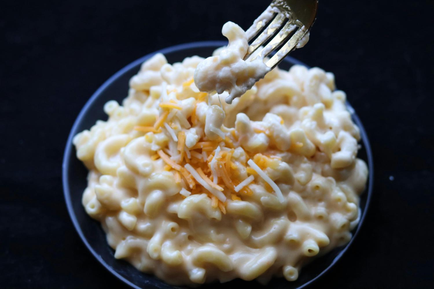 A bowl of macaroni and cheese with shredded cheese on top is shown on a black surface, with a fork lifting some of the macaroni up. The cheese is creamy and dripping from the fork.
