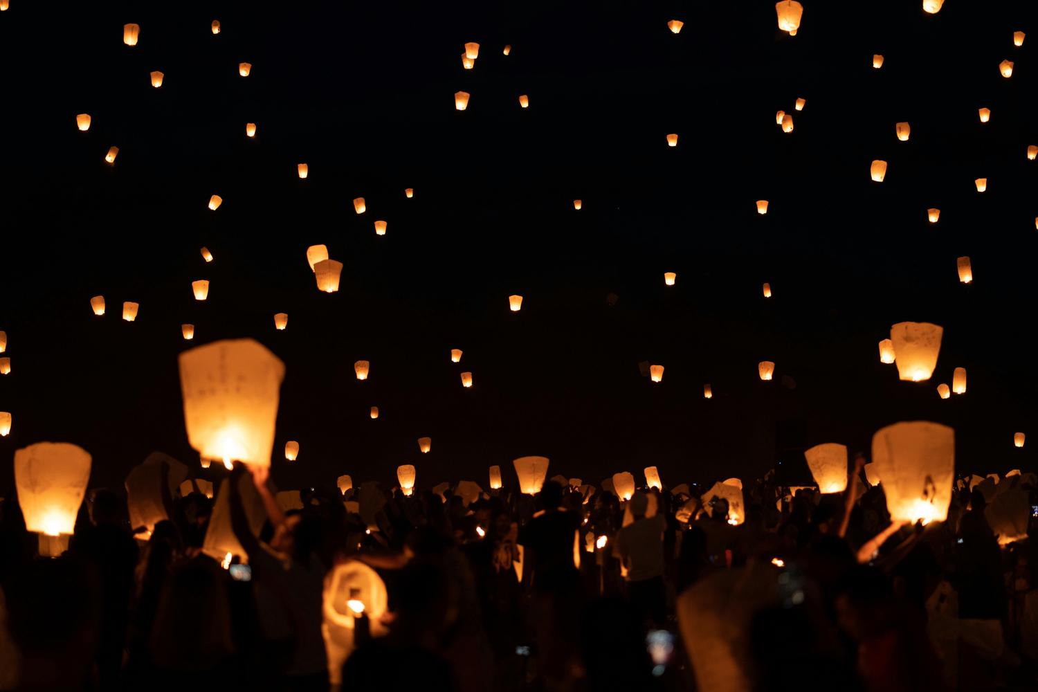 A large crowd releases many illuminated sky lanterns into the dark night, creating a magical and festive atmosphere. The lanterns fill the sky and are visible in the hands of silhouetted figures below.