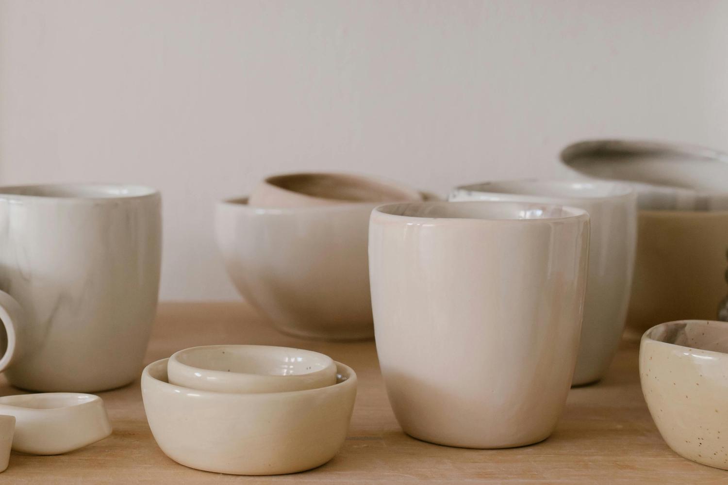 A collection of handmade ceramic bowls and cups in neutral tones sits on a wooden surface against a light background. The vessels vary in size and shape, suggesting a set of pottery still in progress.