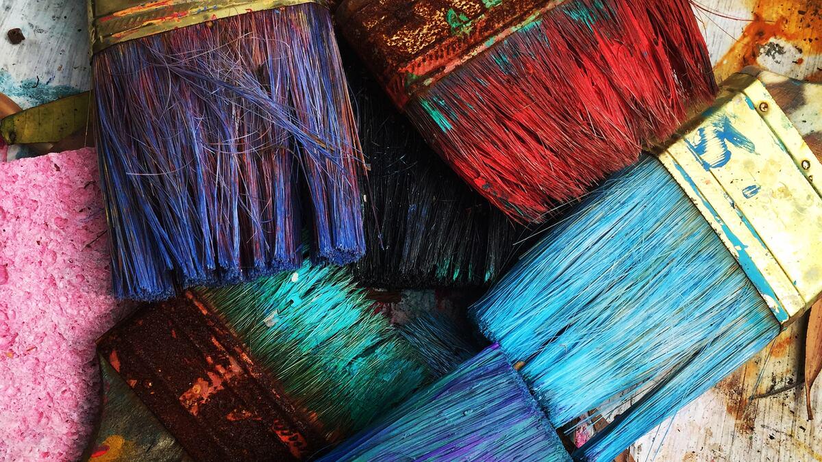 Close-up overhead shot of several paintbrushes with varying colored bristles, including blue, red, and teal, arranged haphazardly on a surface with paint splatters and a pink sponge. The brushes appear well-used and covered in dried paint.