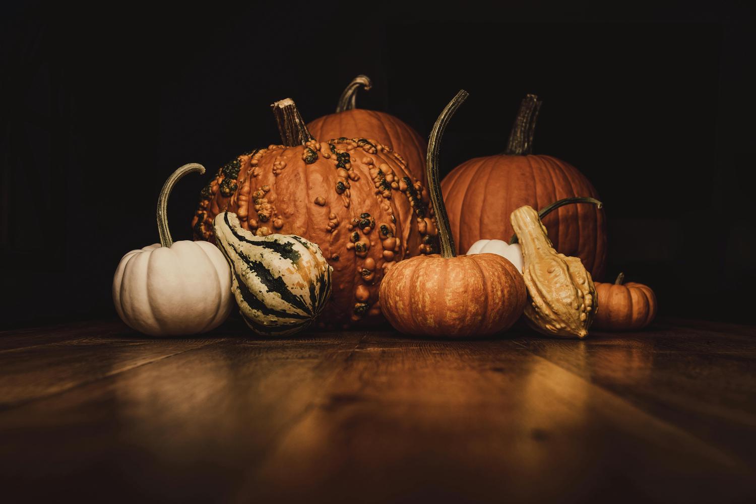 A collection of pumpkins and gourds in varying sizes, colors, and textures sits on a dark wooden surface against a dark background. The pumpkins include orange, white, and striped varieties, some with bumpy textures.