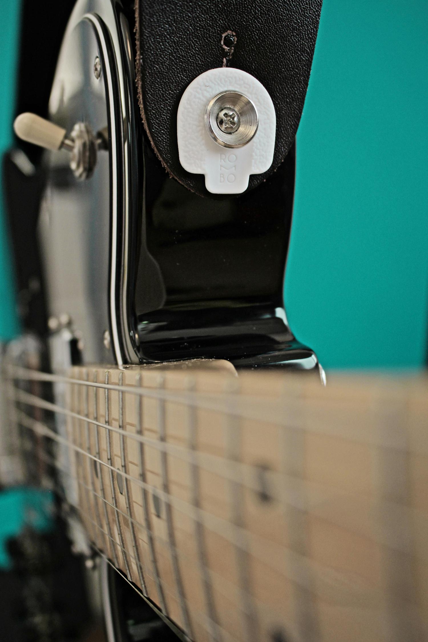 Close-up of an electric guitar neck and body with a black leather strap attached using a white Rombo strap lock system. The background is teal.