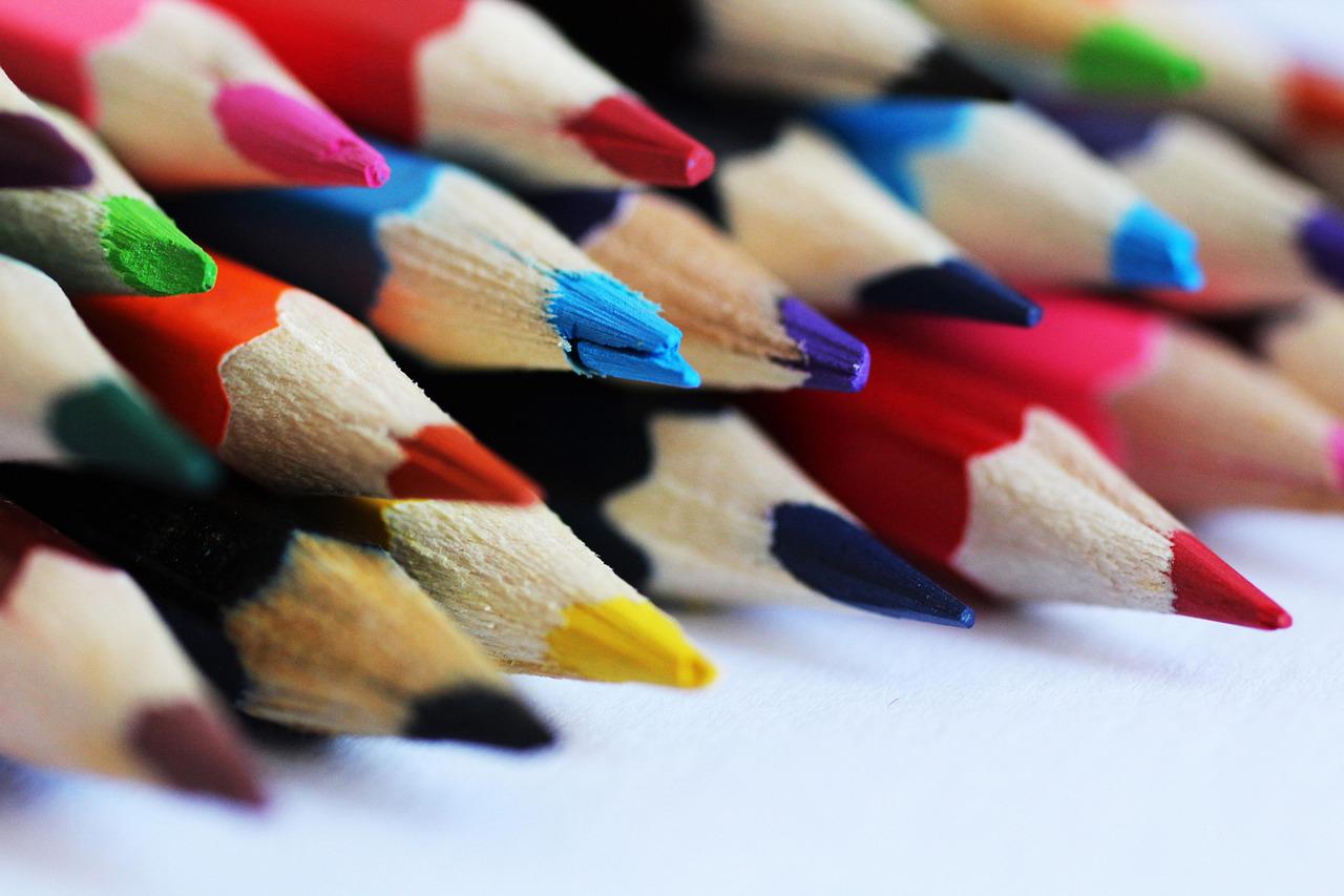 Close-up of a row of colored pencils with points facing right, showing colors like red, pink, blue, green, and yellow. The wood grain of the pencils is visible.