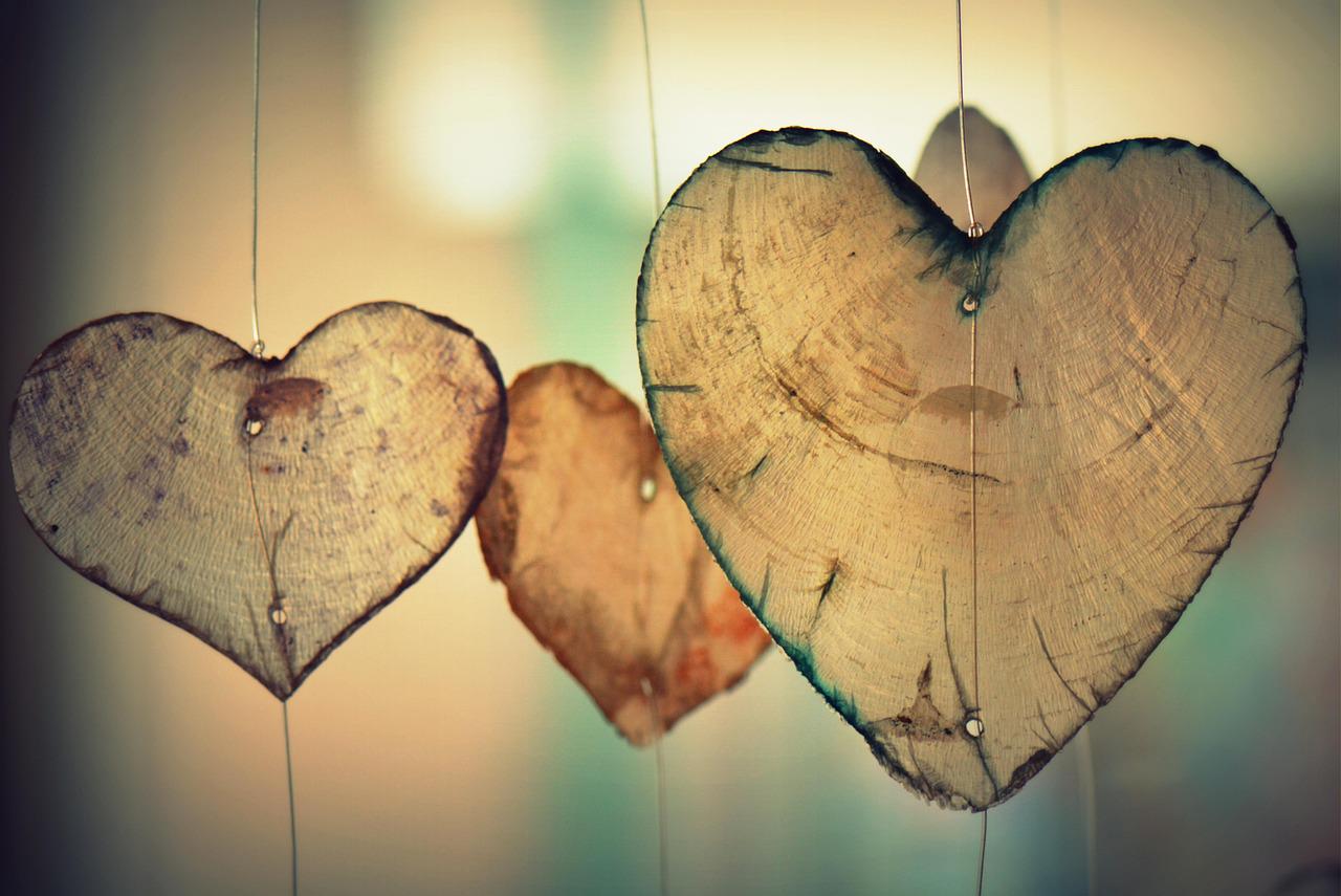 Close-up of three wooden heart-shaped ornaments hanging from thin strings. The hearts show wood grain details and small holes, creating a rustic and romantic aesthetic.
