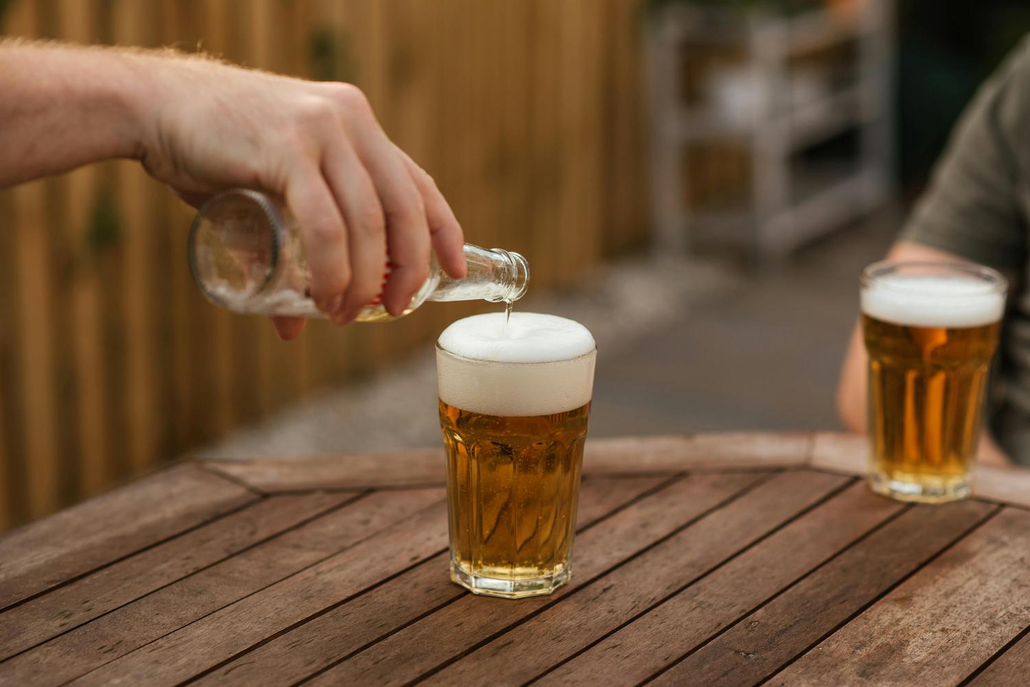 A hand pours beer from a bottle into a glass, creating a frothy head; another filled glass sits on the wooden table.