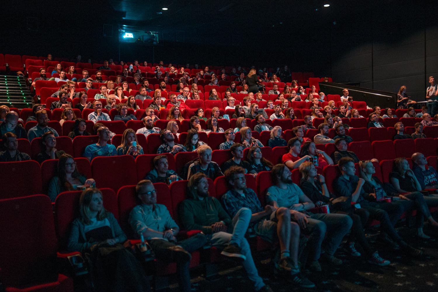 A wide view of a darkened movie theater filled with an audience seated in red chairs, looking toward the screen. A few people are standing at the top rows.