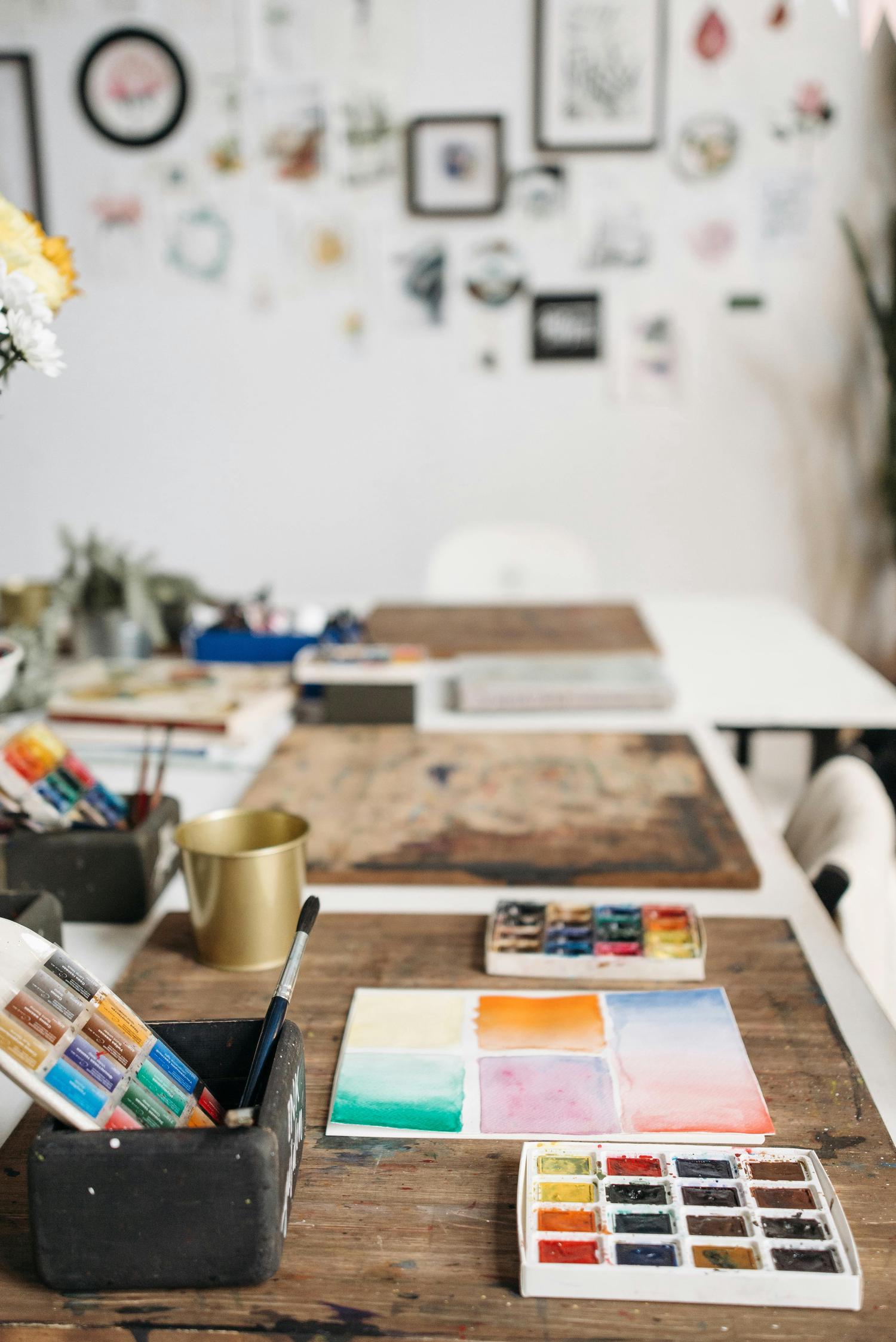 A close-up view of an artist's workspace featuring watercolor paints, brushes in a container, and a watercolor test sheet with colorful blocks. The background shows a long white table with art supplies and a wall decorated with various pictures and artworks.
