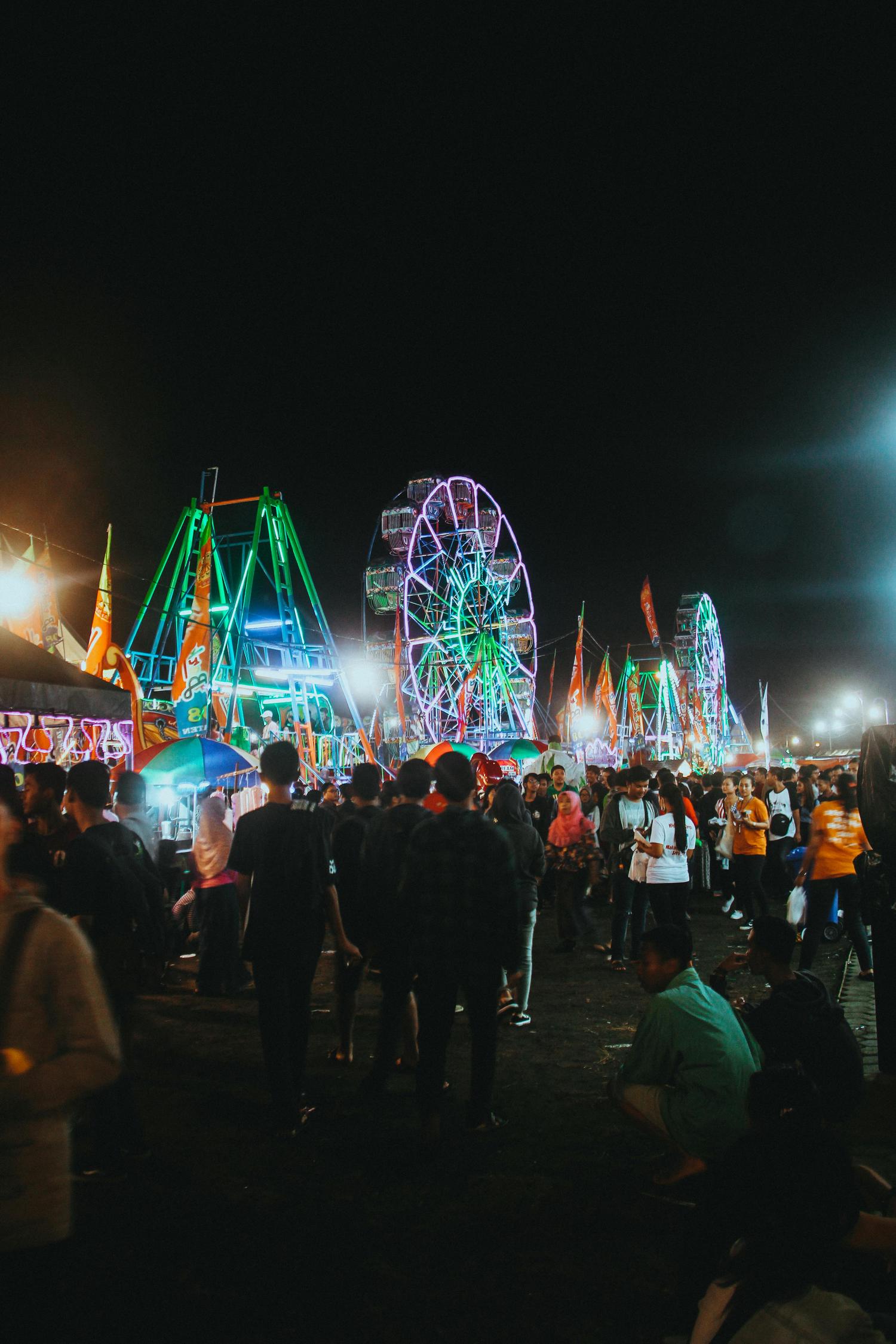 A nighttime scene shows a crowded fair or festival, with several brightly lit carnival rides like Ferris wheels visible in the background. People are milling around, some standing and some sitting, creating a sense of lively activity.