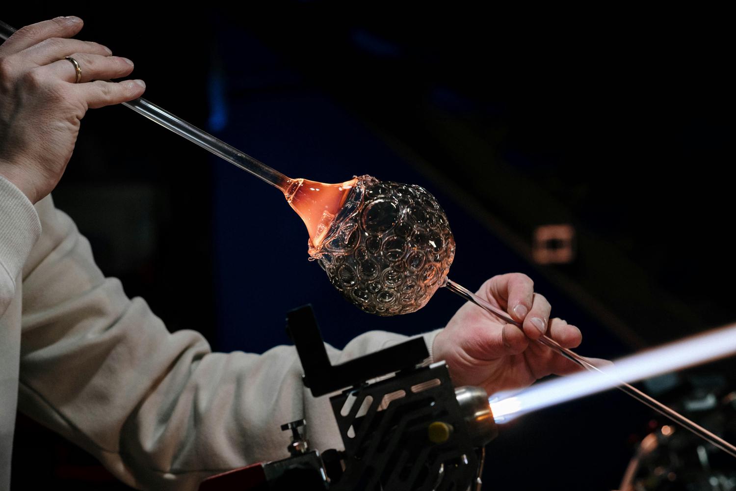 A glassblower shapes molten glass attached to a blowpipe while another tool is heated with a torch. The glass has many circular indentations and glows red near the attachment point.