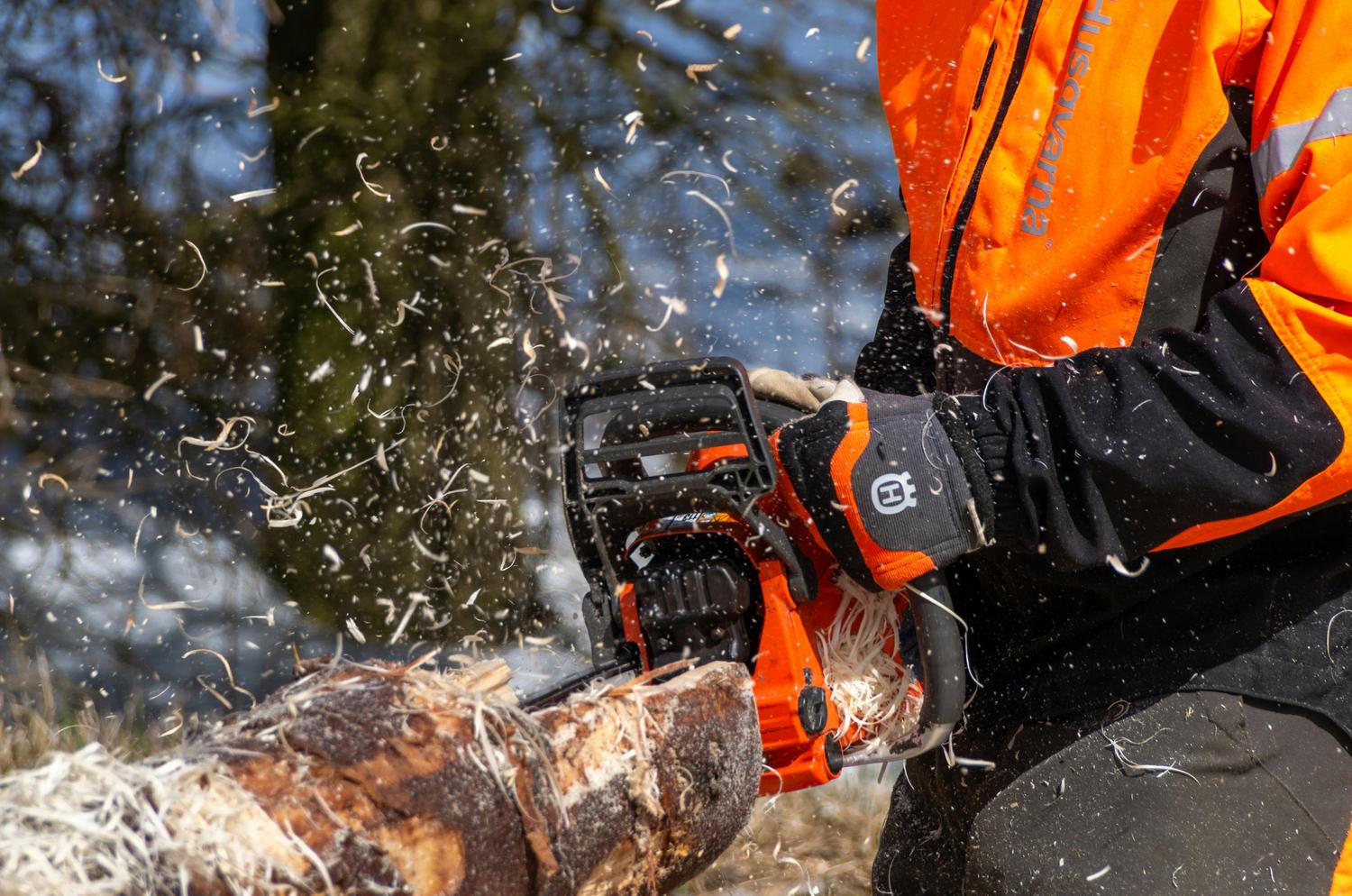 A person wearing safety gear uses a Husqvarna chainsaw to cut a log, sending wood shavings flying. The worker's bright orange jacket is visible, along with a gloved hand operating the chainsaw.