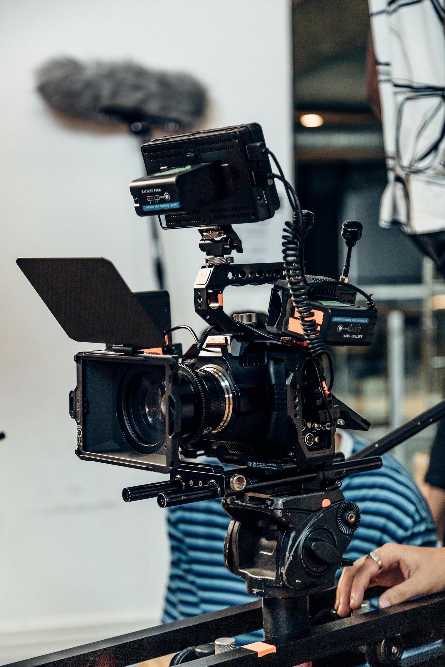 A professional film camera with accessories like a matte box, battery pack, and microphone is mounted on a dolly track. A hand with a ring is visible, adjusting the dolly.