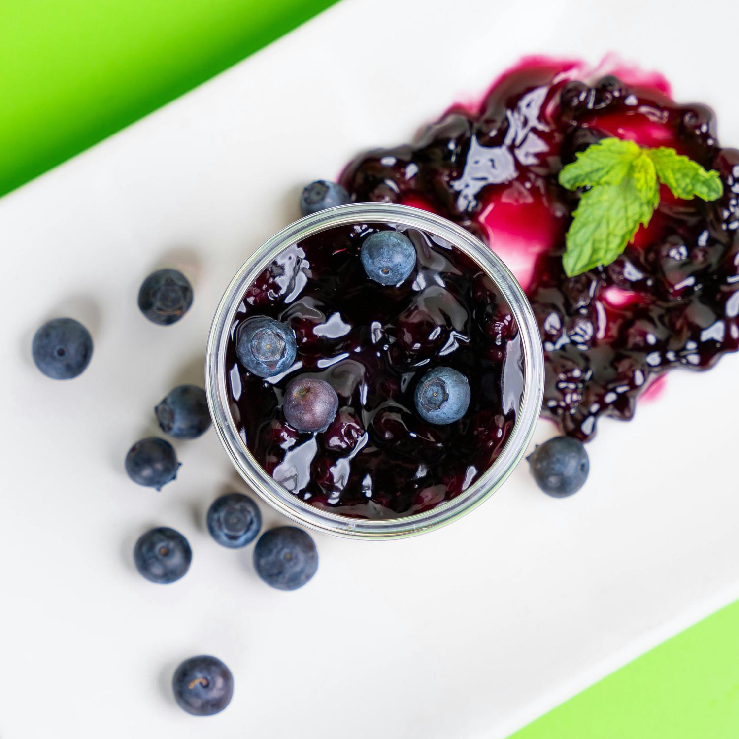 Overhead shot of a small glass container filled with blueberry sauce and scattered blueberries, next to a spoonful of blueberry sauce garnished with mint on a white plate, set against a green background.