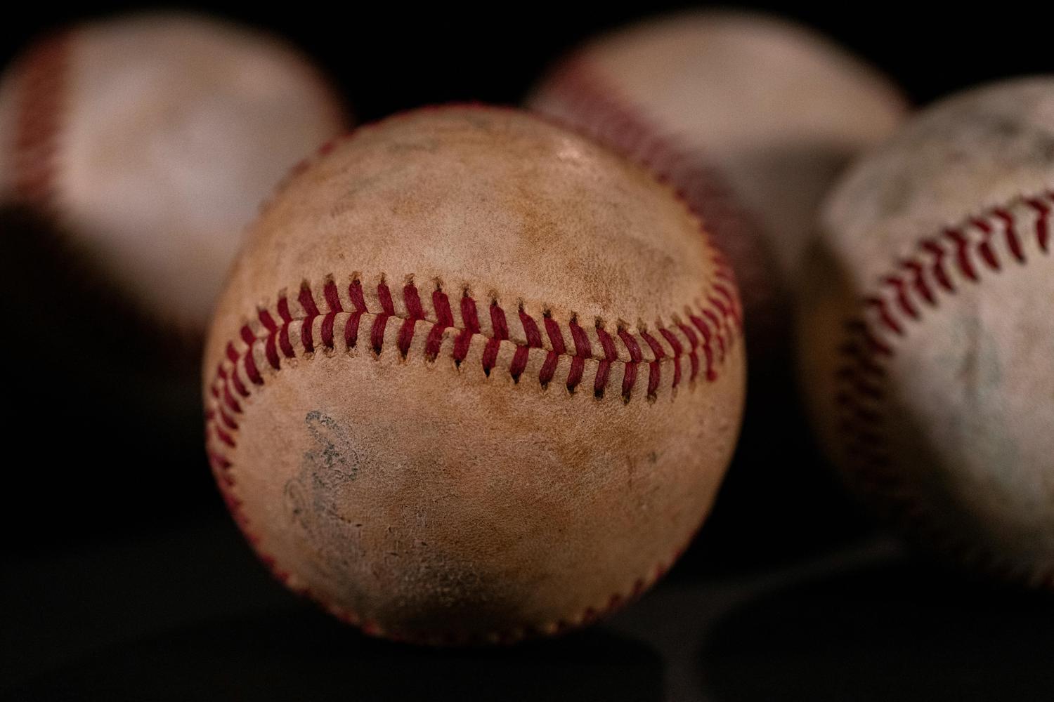 Close-up view of several baseballs, one in sharp focus in the foreground with red stitching, against a dark, blurred background. The baseballs appear well-used and textured.