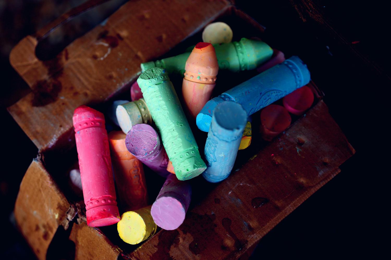 A close-up shot depicts a cardboard box overflowing with vibrantly colored chalk pastels in hues of pink, purple, yellow, blue, orange, and green. The chalk pieces vary in size and shape, filling the box to the brim.