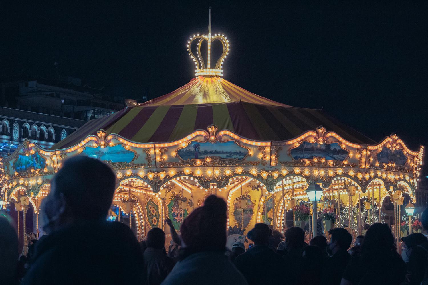 A brightly lit carousel stands at night, topped with a striped tent and a crown of lights. Silhouetted figures of people stand in the foreground, looking at the carousel.