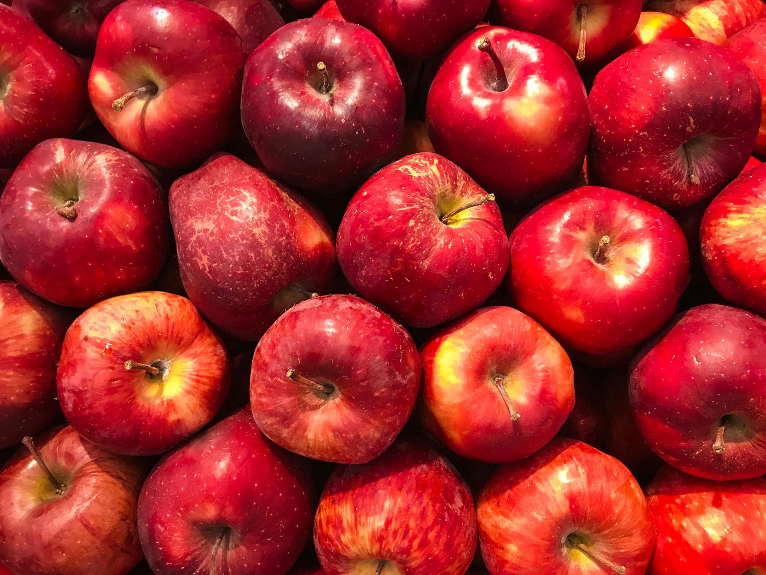 A close-up of a heap of red apples, filling the frame, with some stems visible. The apples have varying shades of red with subtle yellow and white speckles.