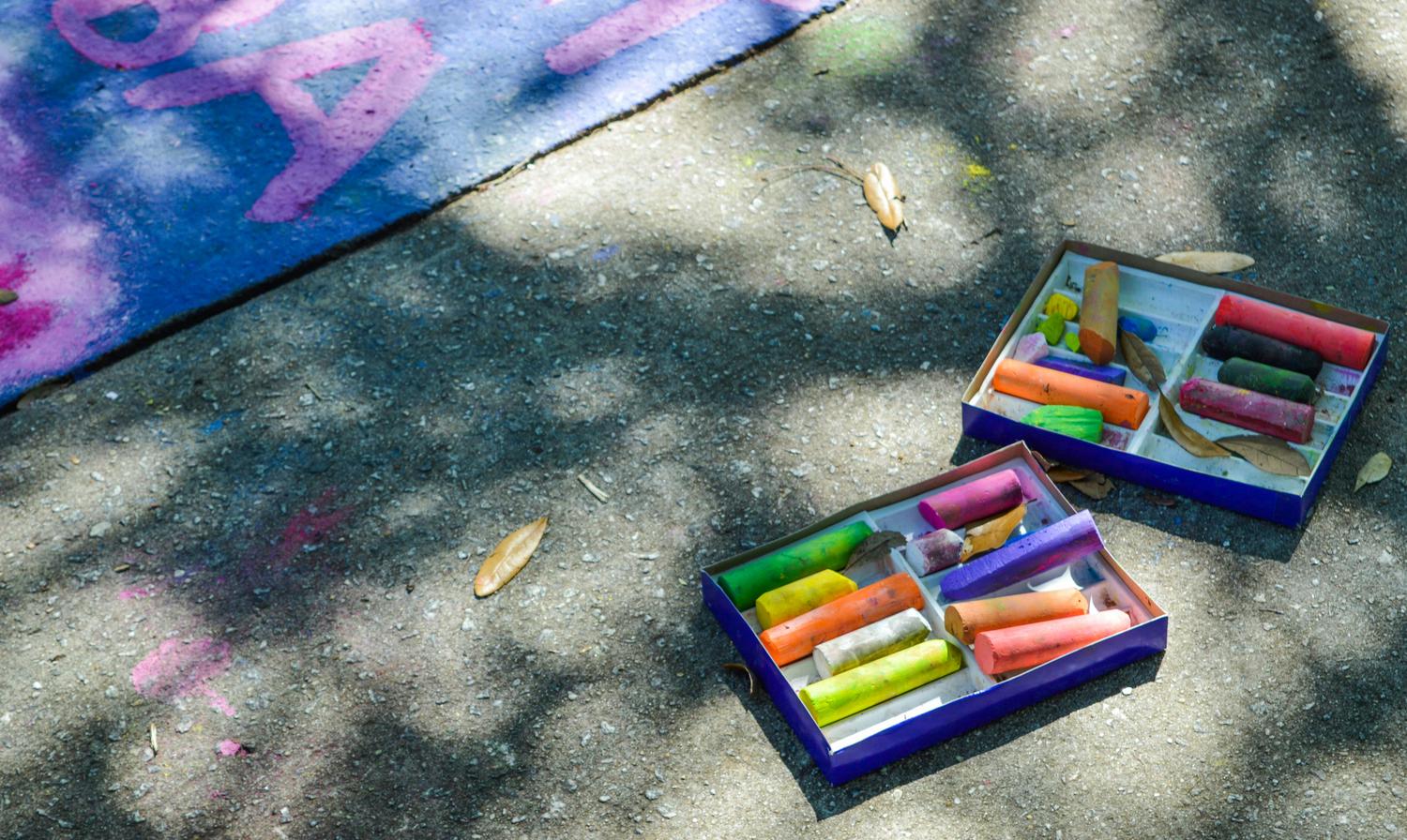 Two boxes of colorful sidewalk chalk sit on a concrete surface next to a patch of blue and pink chalk art, dappled with sunlight and shadows. Several small brown leaves are scattered nearby.