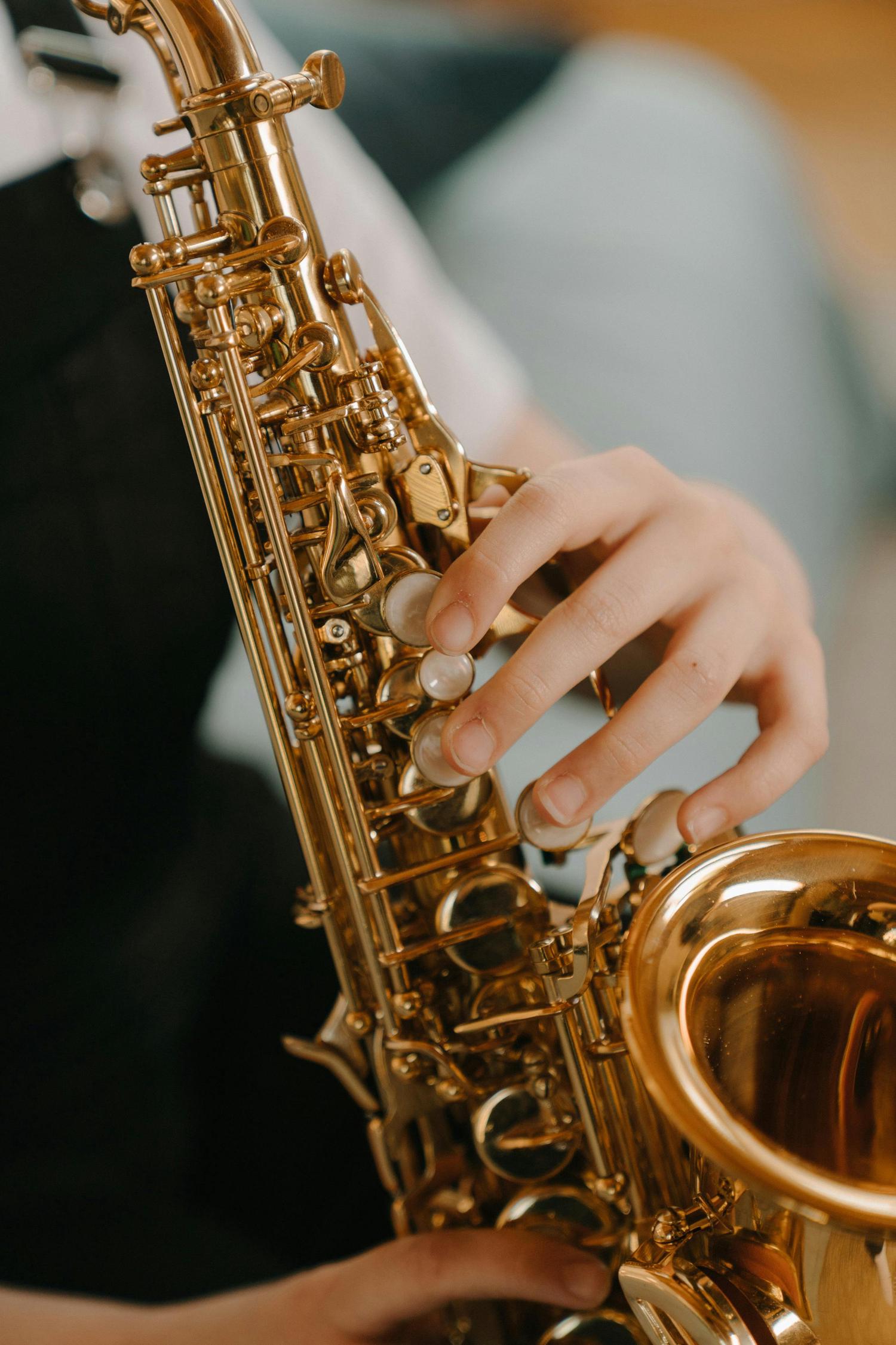 A person's hand is pressing the keys of a bright gold saxophone. The instrument is vertical and fills the center of the frame, with a blurred figure in the background.