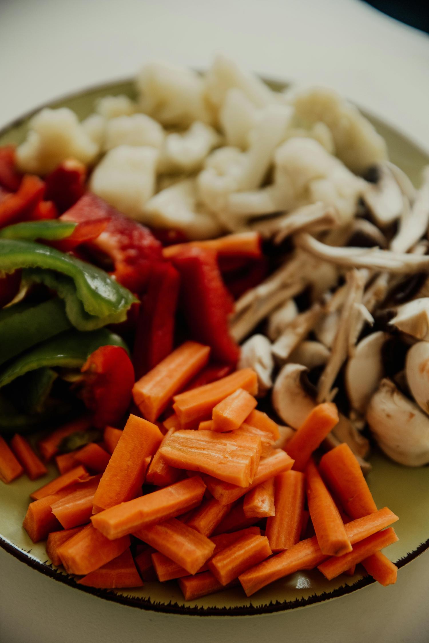 A plate contains neatly arranged, chopped vegetables: bright orange carrots, green and red bell peppers, white cauliflower florets, and sliced white mushrooms. The vegetables appear freshly cut and ready for cooking.
