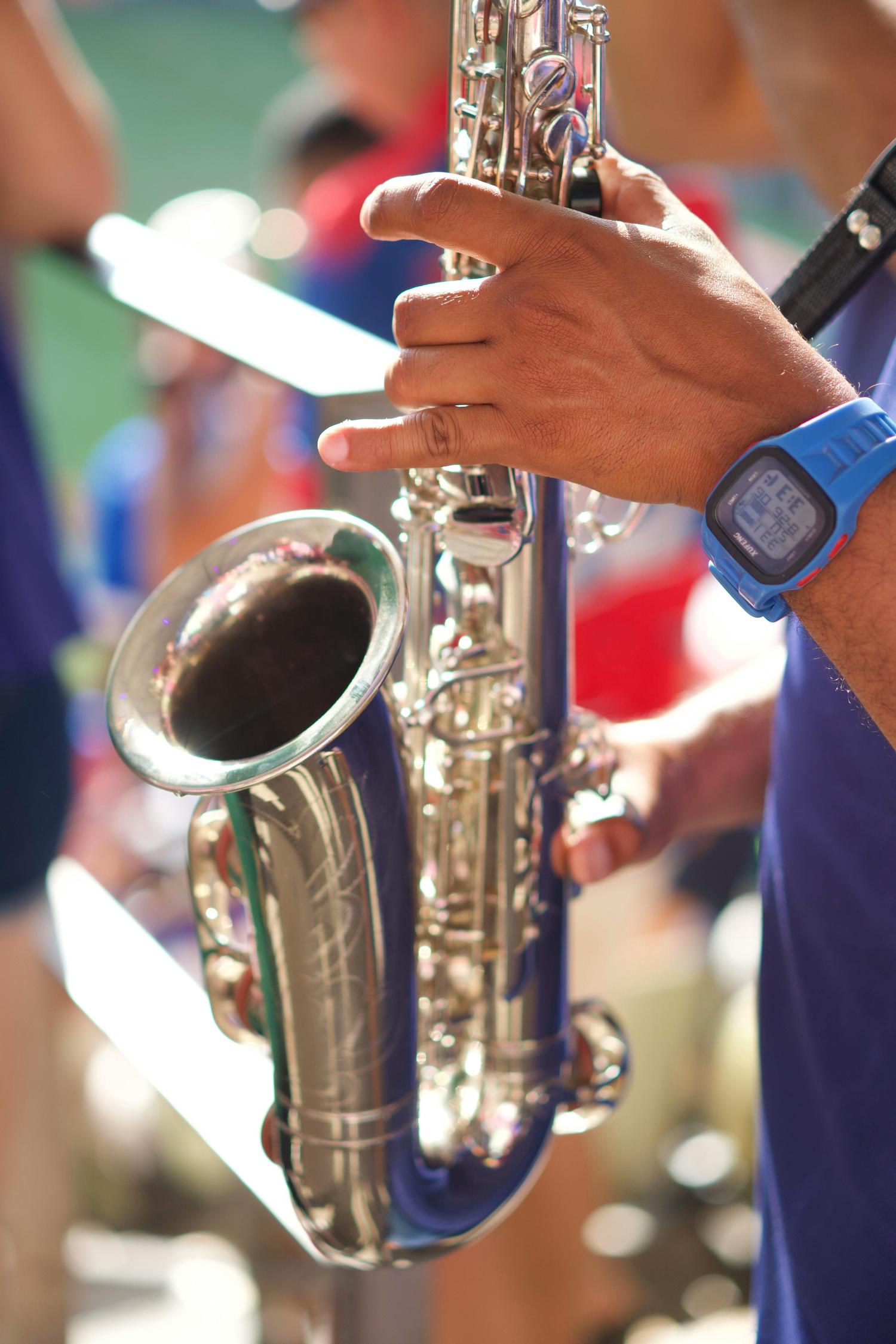 Close-up of hands holding a silver saxophone, with a blue digital watch visible on the player's wrist. The musician is wearing a purple shirt, and other people are visible in the blurred background.